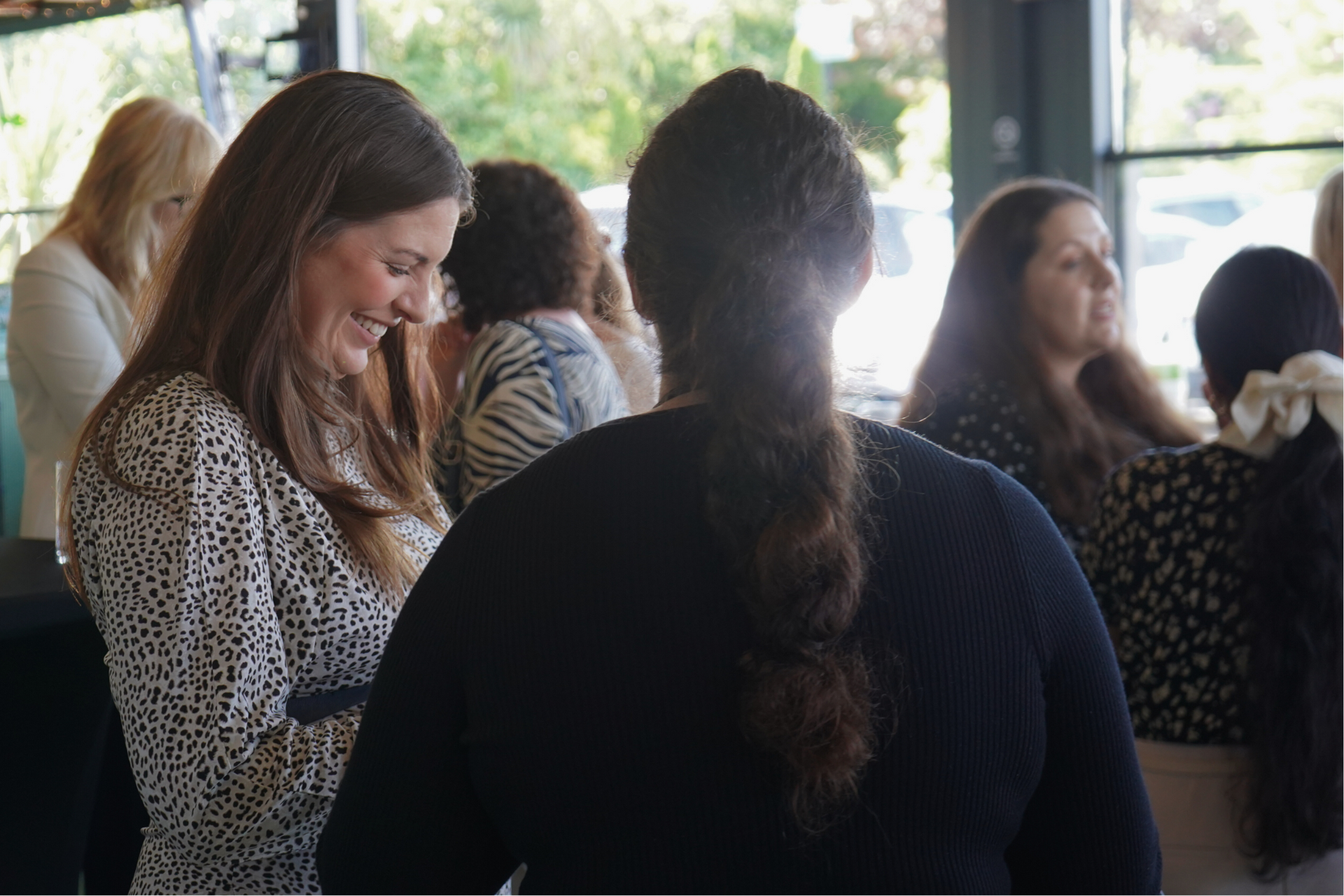 People gathered indoors, chatting and smiling near large windows.