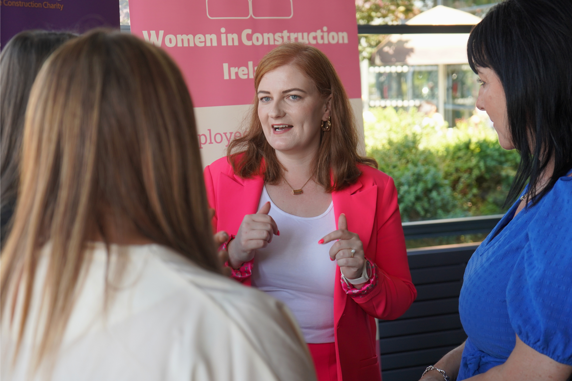 Three women converse outdoors; one in a red jacket gestures while speaking in front of a banner.
