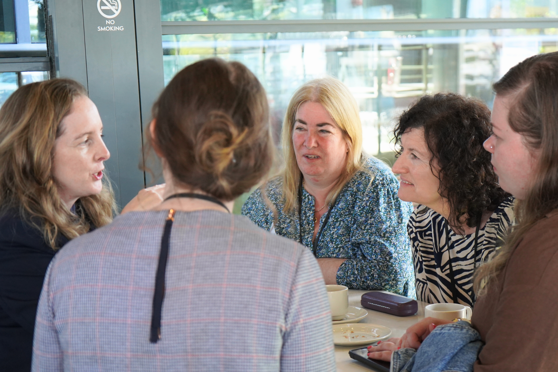 Group of people chatting around a table in a bright cafe, with one smiling woman facing the camera.