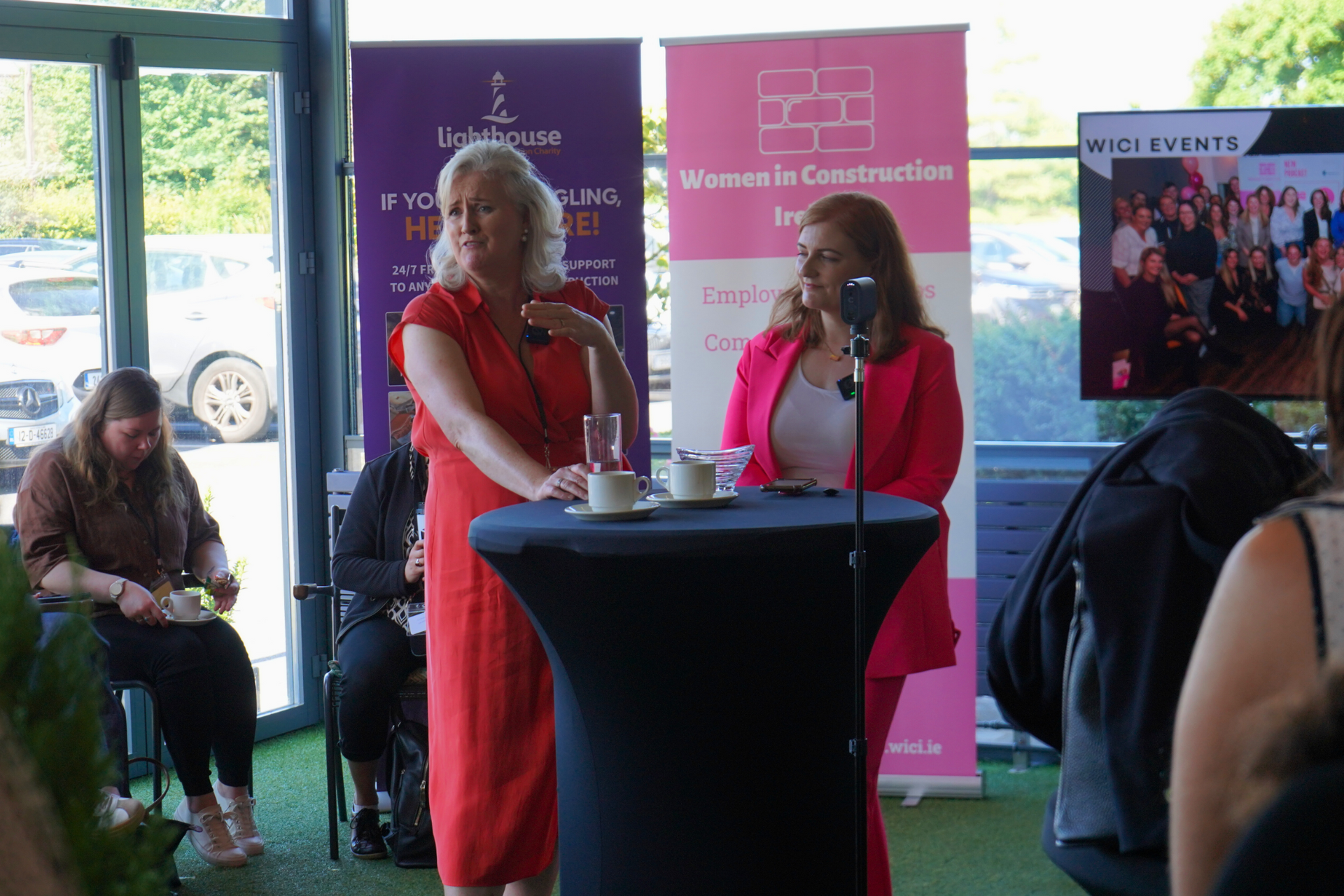 Two women speaking at podiums during an indoor event with banners and audience photos in the background
