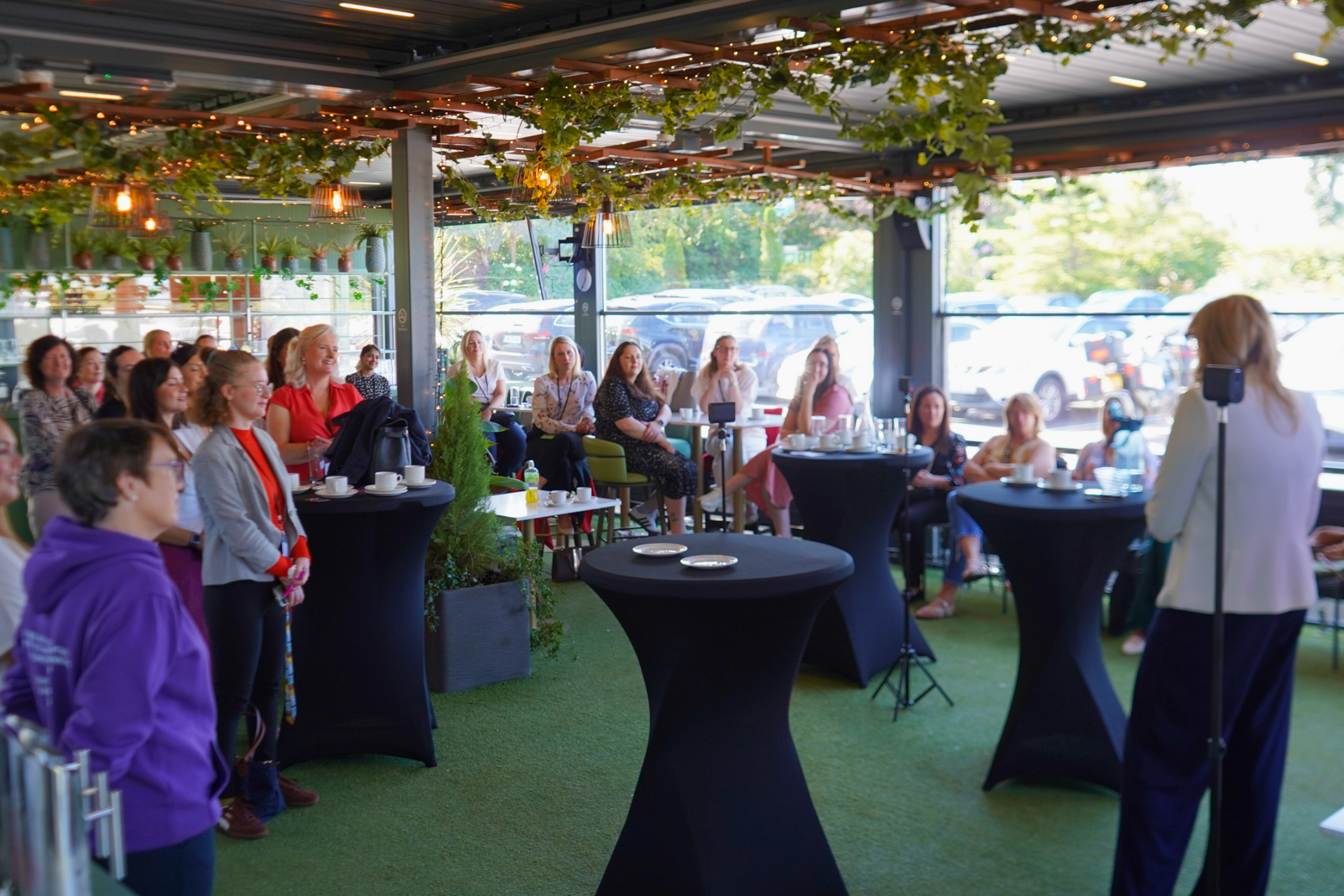 People gathered at a decorated indoor event space, listening to a speaker at a microphone.