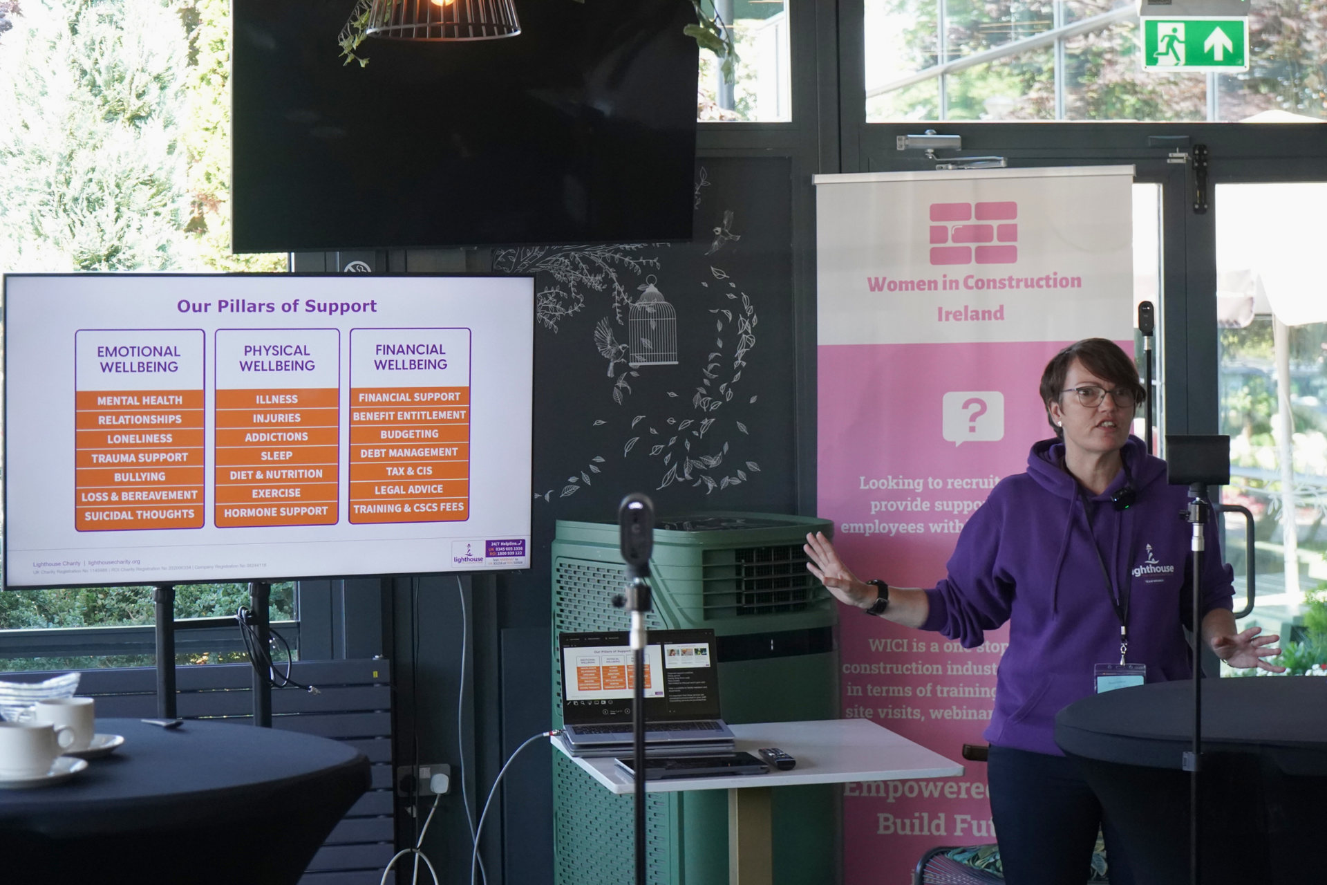 Person in a purple shirt presenting beside slides and a sign at an indoor event booth
