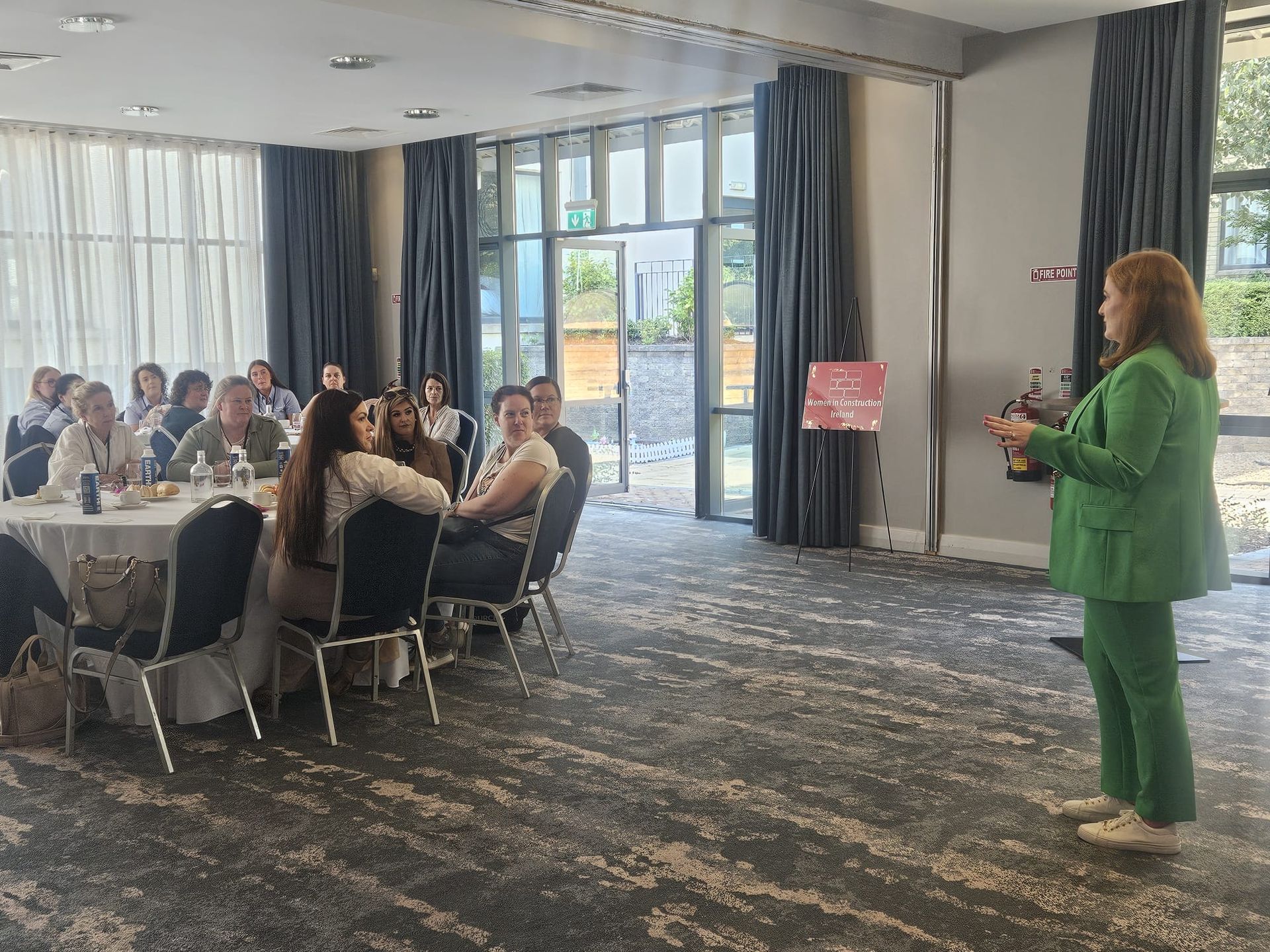 Meeting room with seated audience and a woman in a green suit presenting near the windows