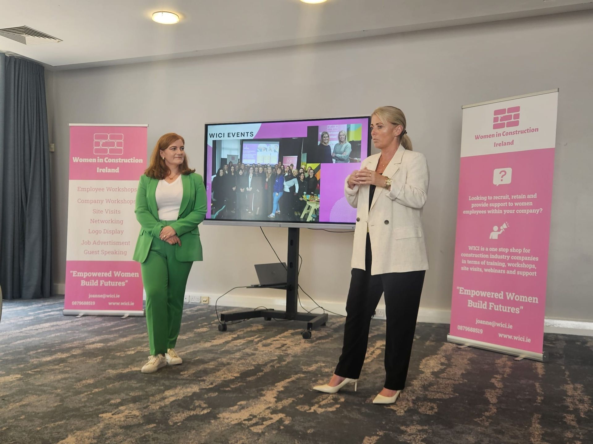 Two women presenting in a conference room beside a large display and pink branded banners.