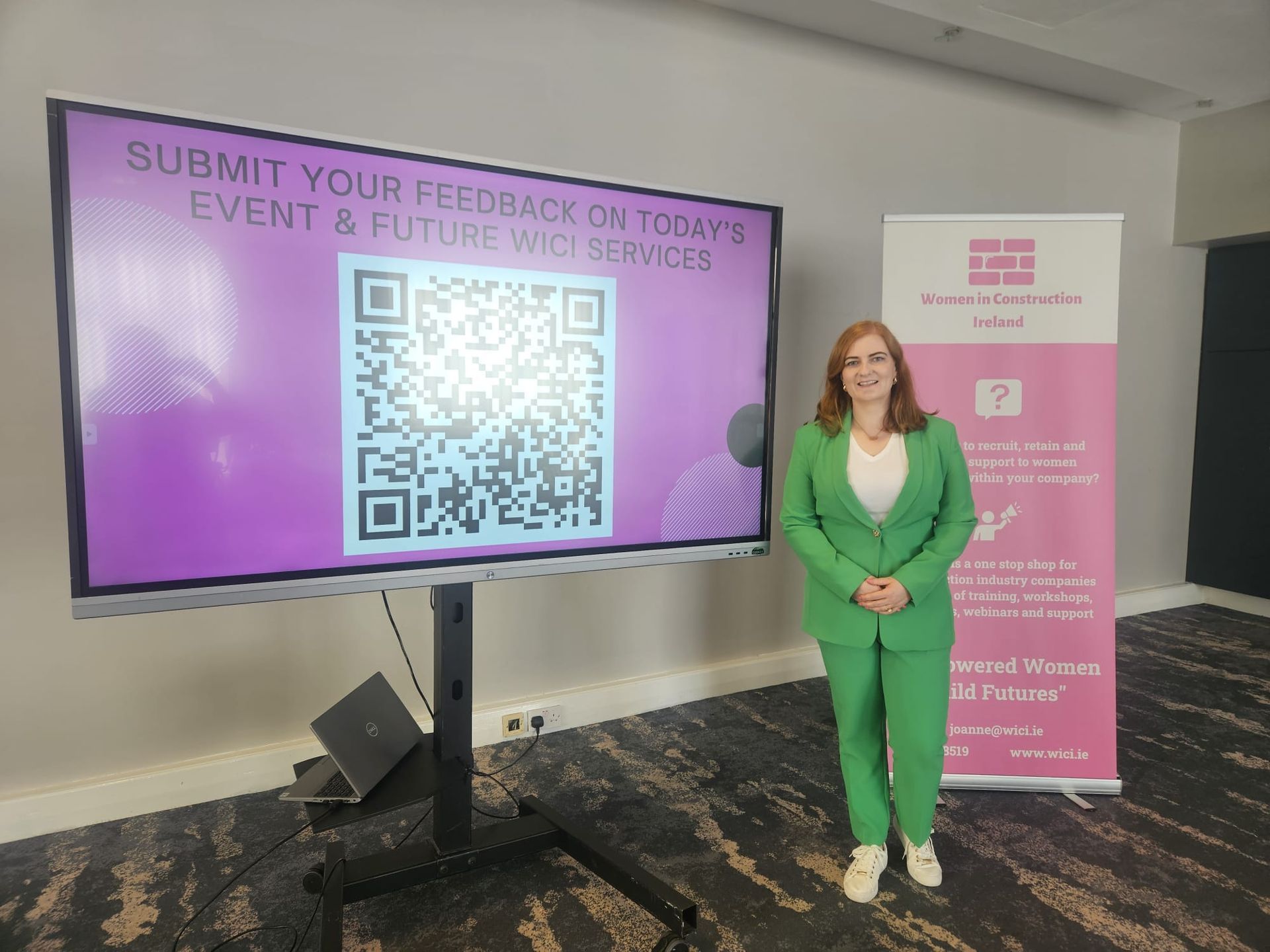 Woman in green suit beside a digital sign and QR code for event feedback, in a lobby setting