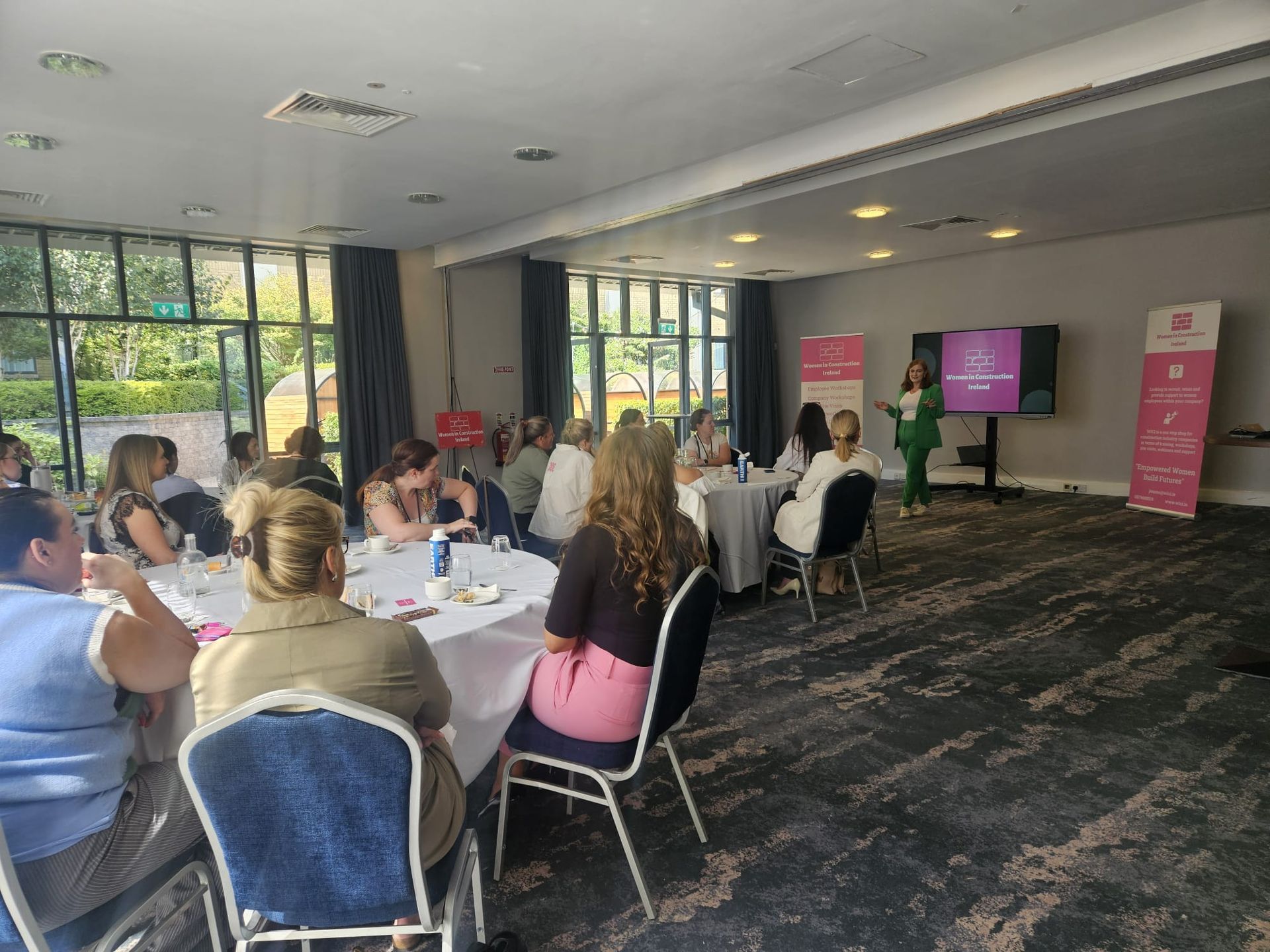 Meeting room with seated attendees watching a presentation beside pink banners and a screen.