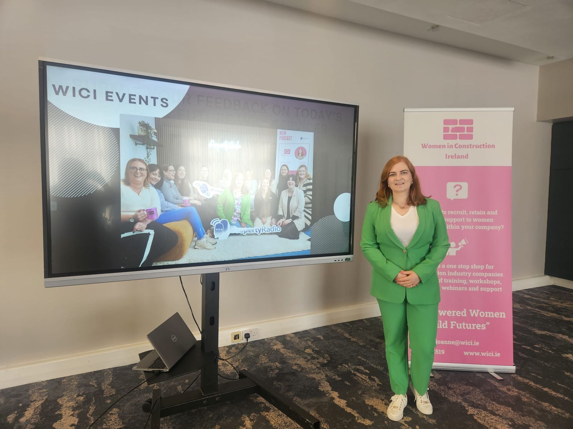 Woman in a green suit stands beside a presentation screen and event banner in a conference room.