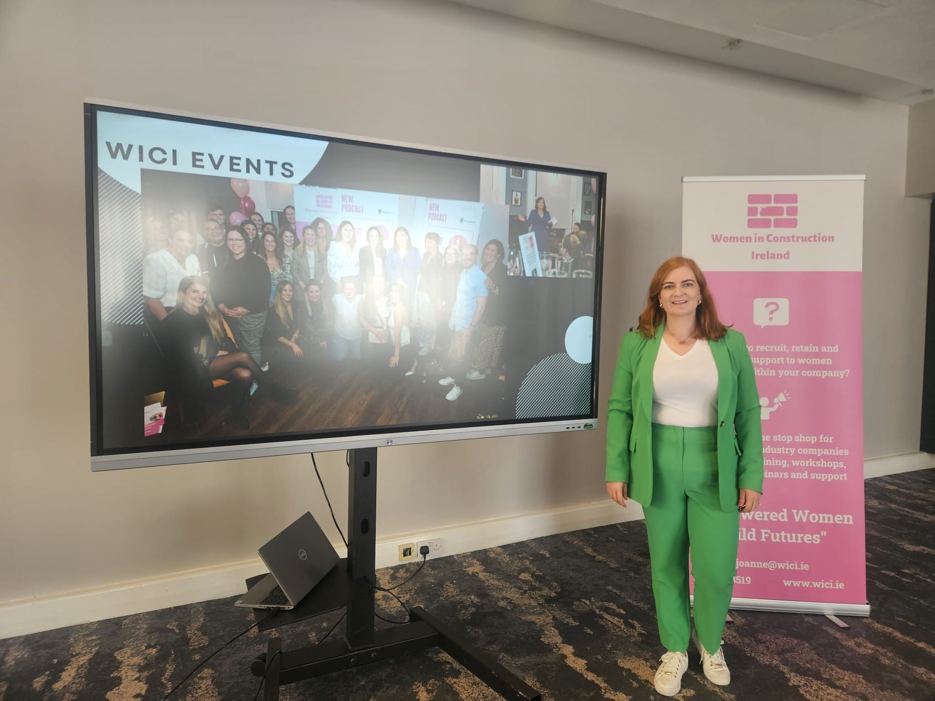 Woman in green suit stands beside a screen showing a group event photo and a pink event banner.