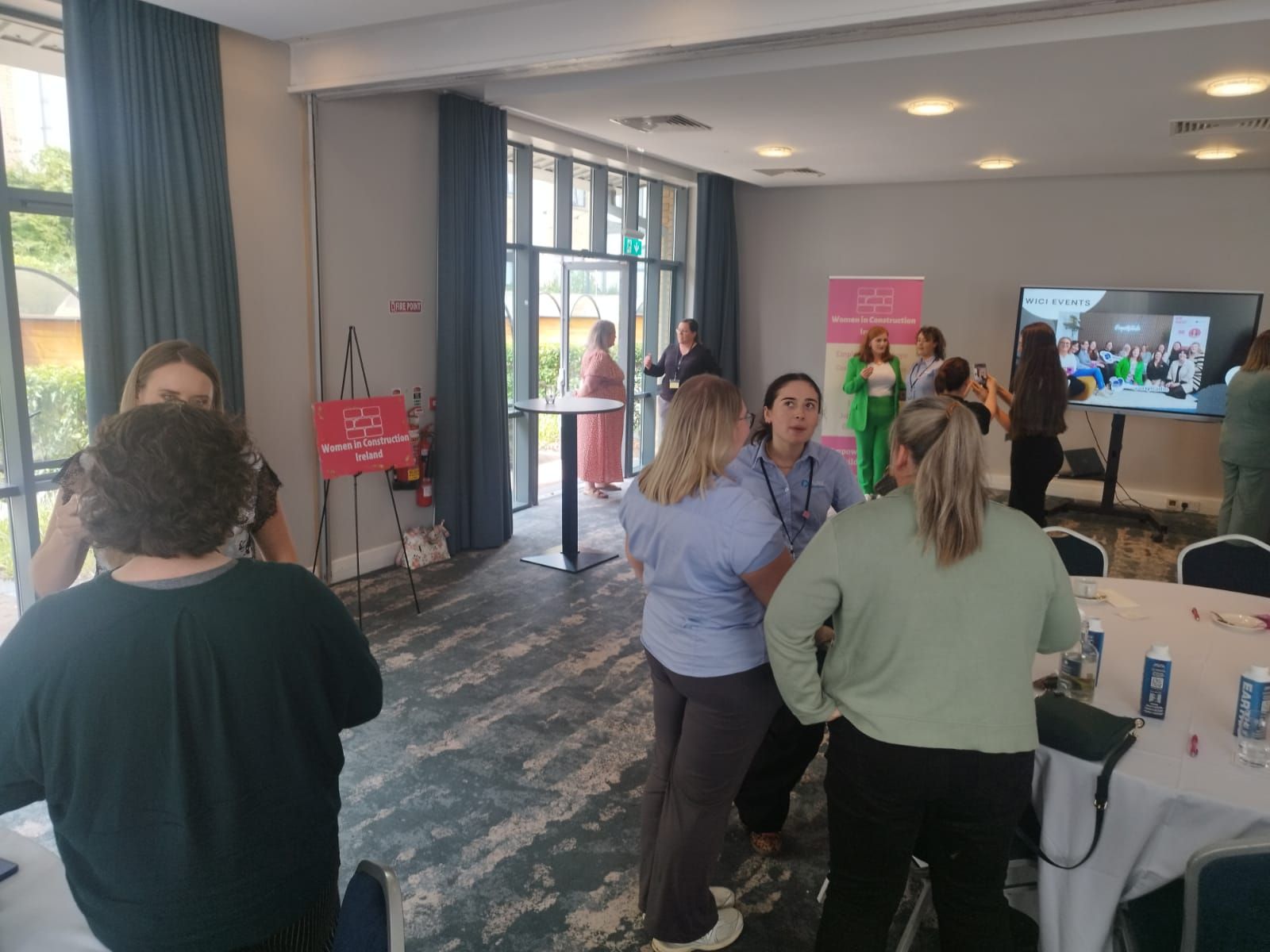 People mingling in a bright room at an indoor event with tables, posters, and a TV screen.
