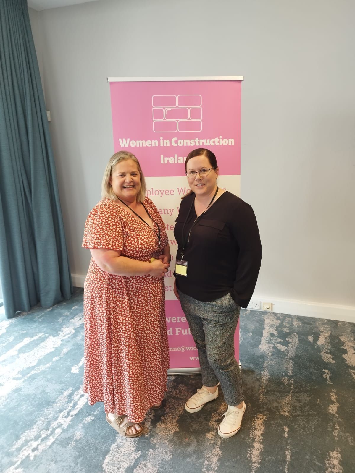 Two women posing beside a pink “Women in Construction” banner in a conference room.