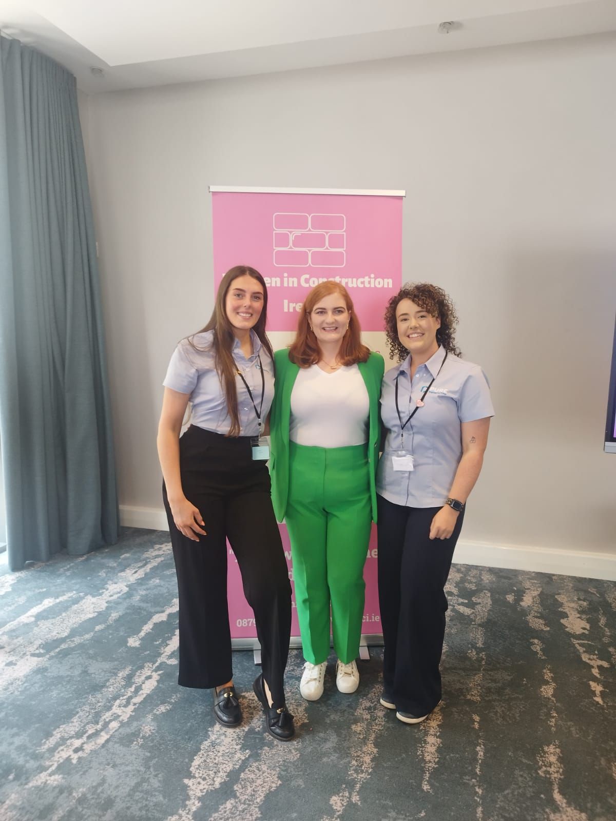 Three people standing together at a conference, smiling in front of a pink event banner.