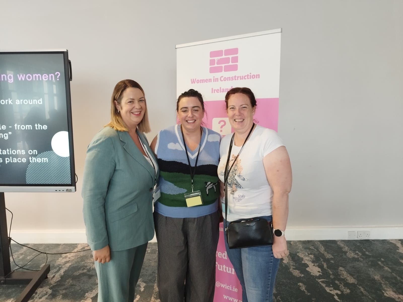 Three people posing indoors beside a pink event banner, smiling at the camera.