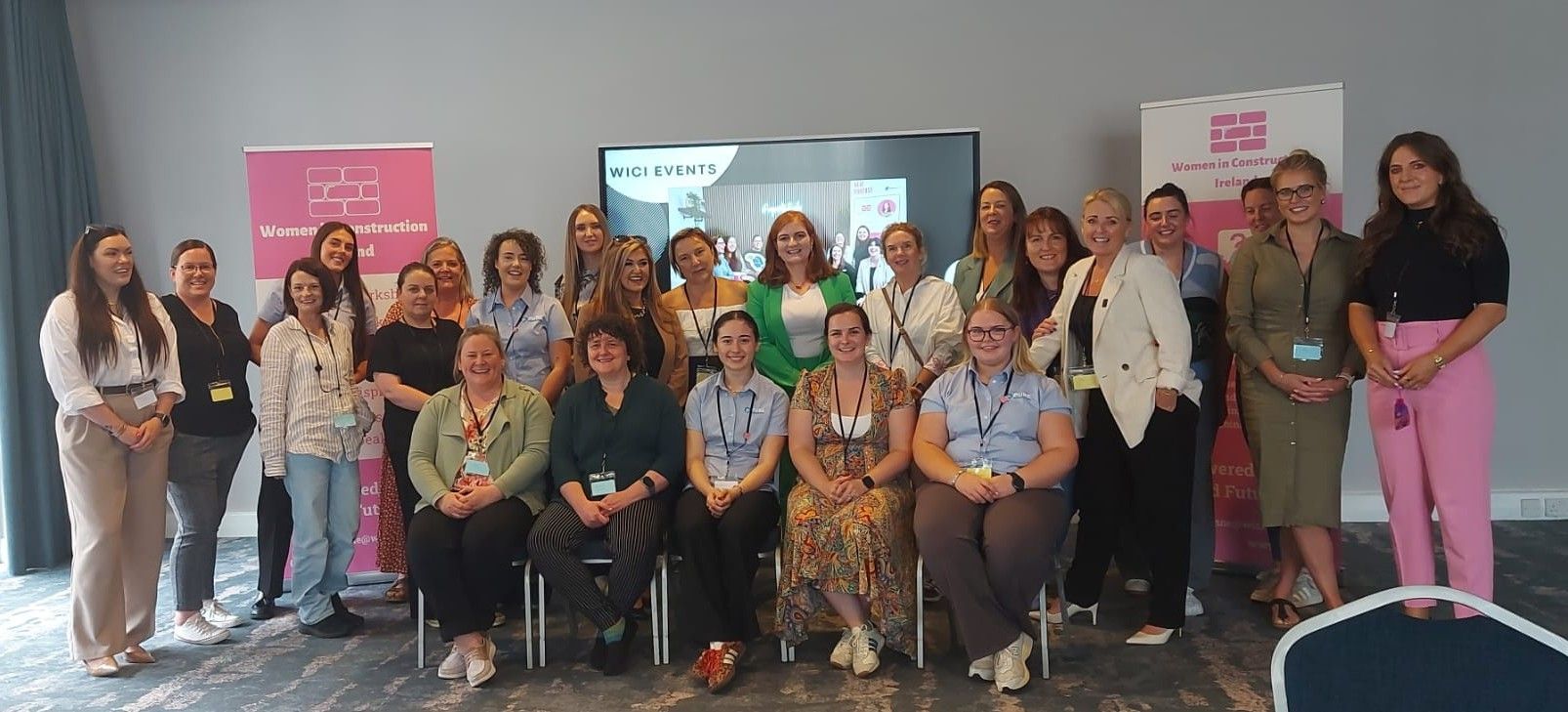 Group of people posing indoors at a conference, with pink banners and a projector screen behind them.