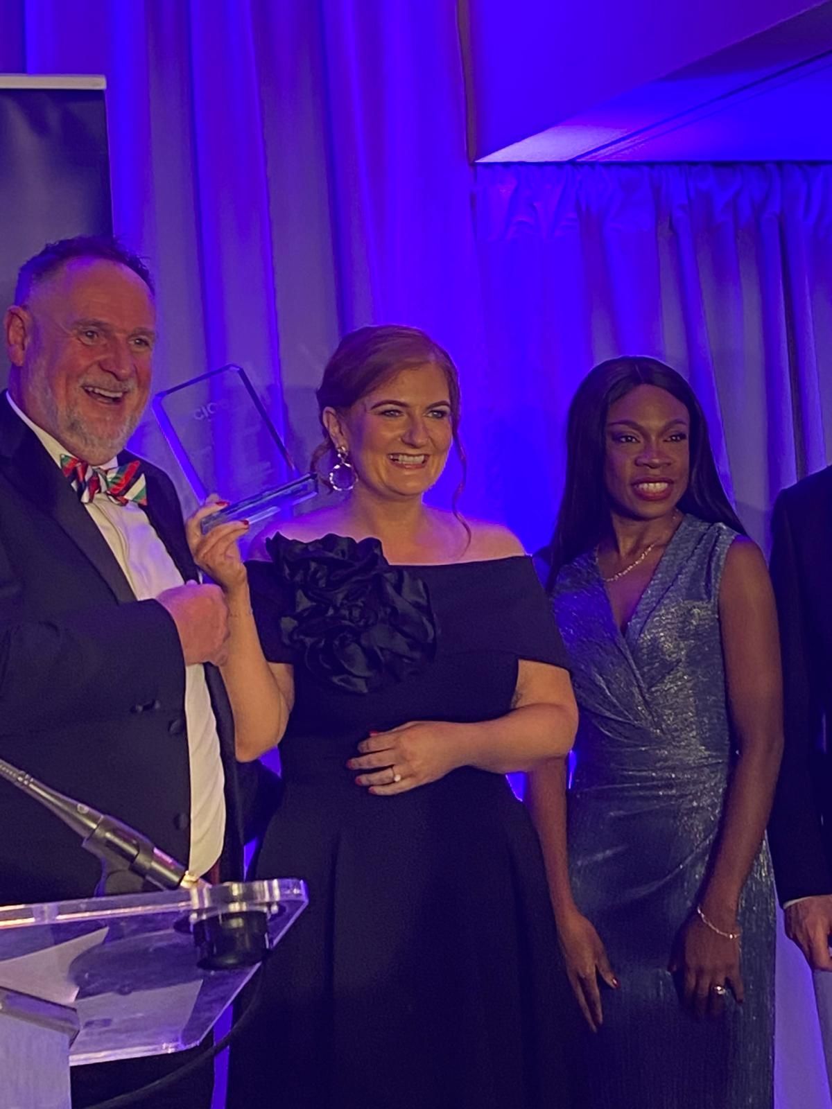 Three people posing at a formal event under blue-purple lighting, one holding a microphone at a podium.