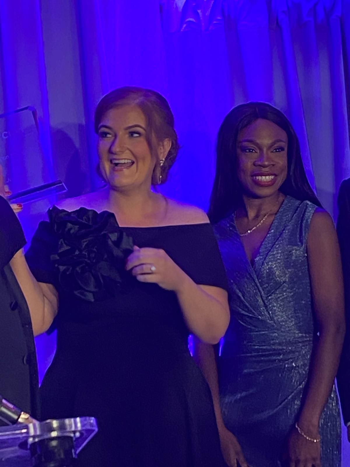 Two women in formal dresses smiling at a blue-lit event, one holding a drink