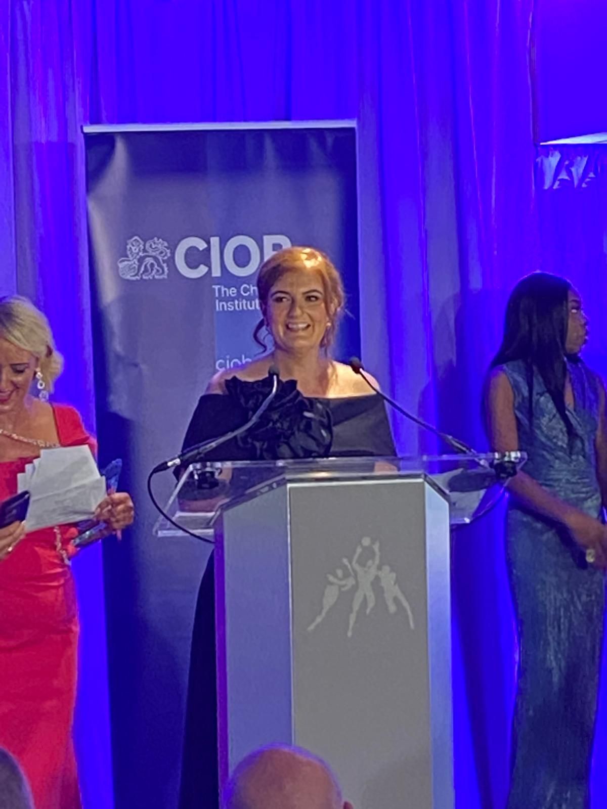 Woman in black dress speaking at a podium on a blue-lit stage, with two people standing nearby.