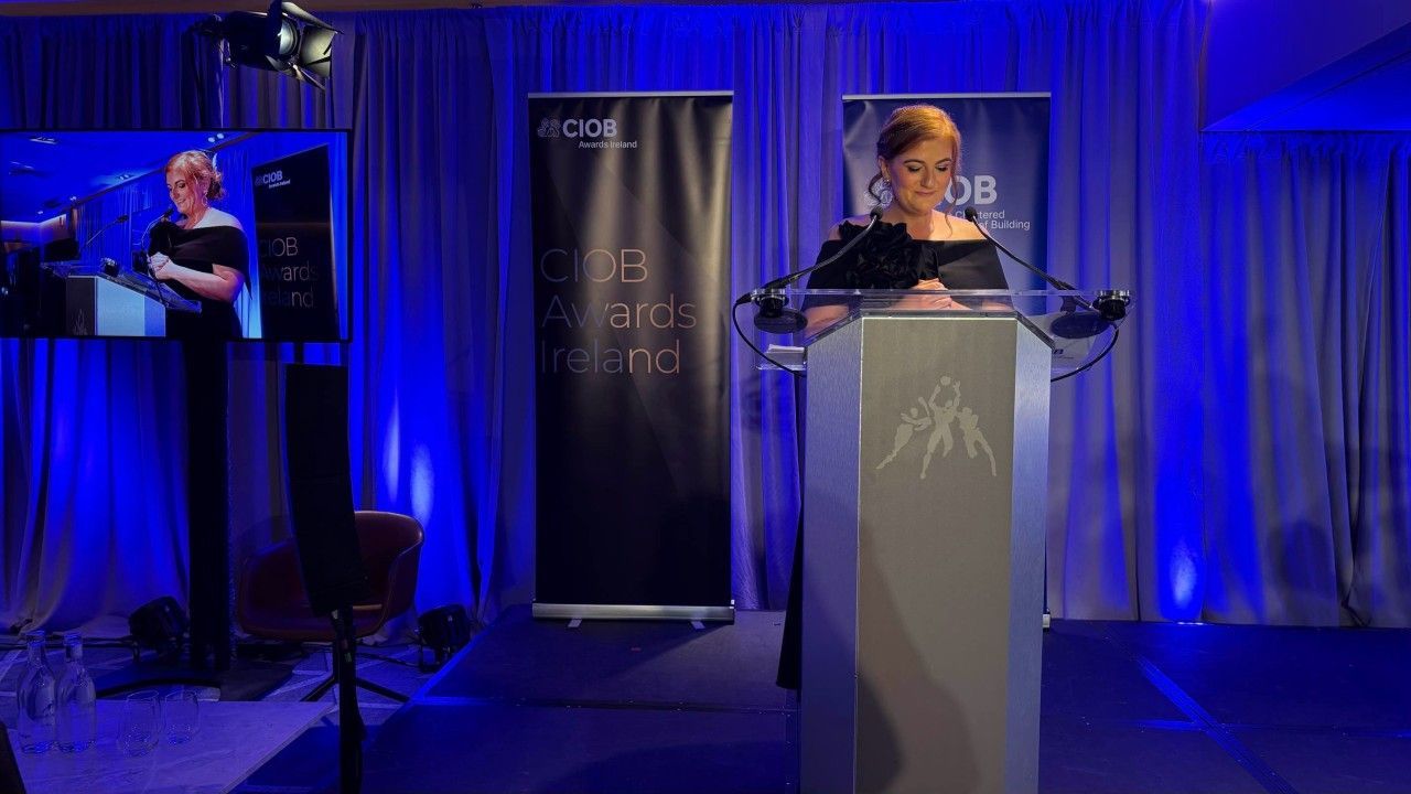 Speaker at podium in blue-lit room, with a signer on a screen at left.
