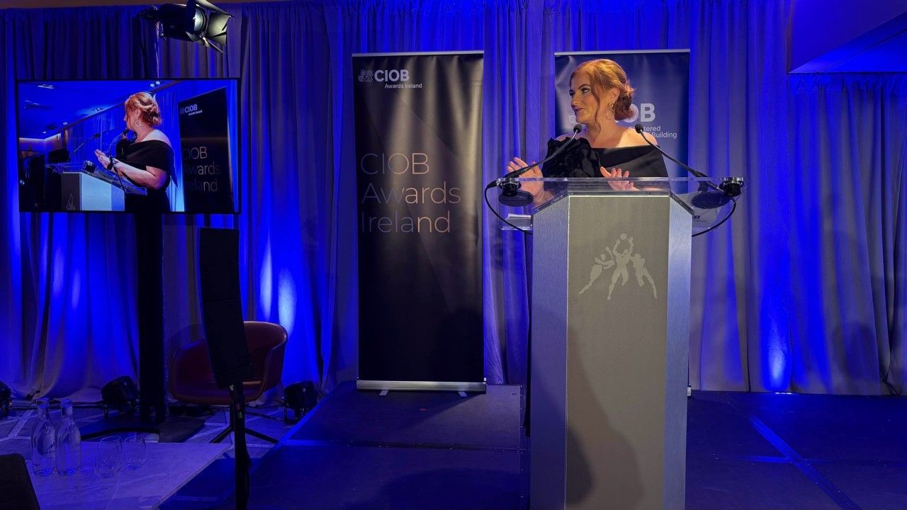 Woman speaking at a podium on a blue-lit stage, with a large screen showing her image beside her.