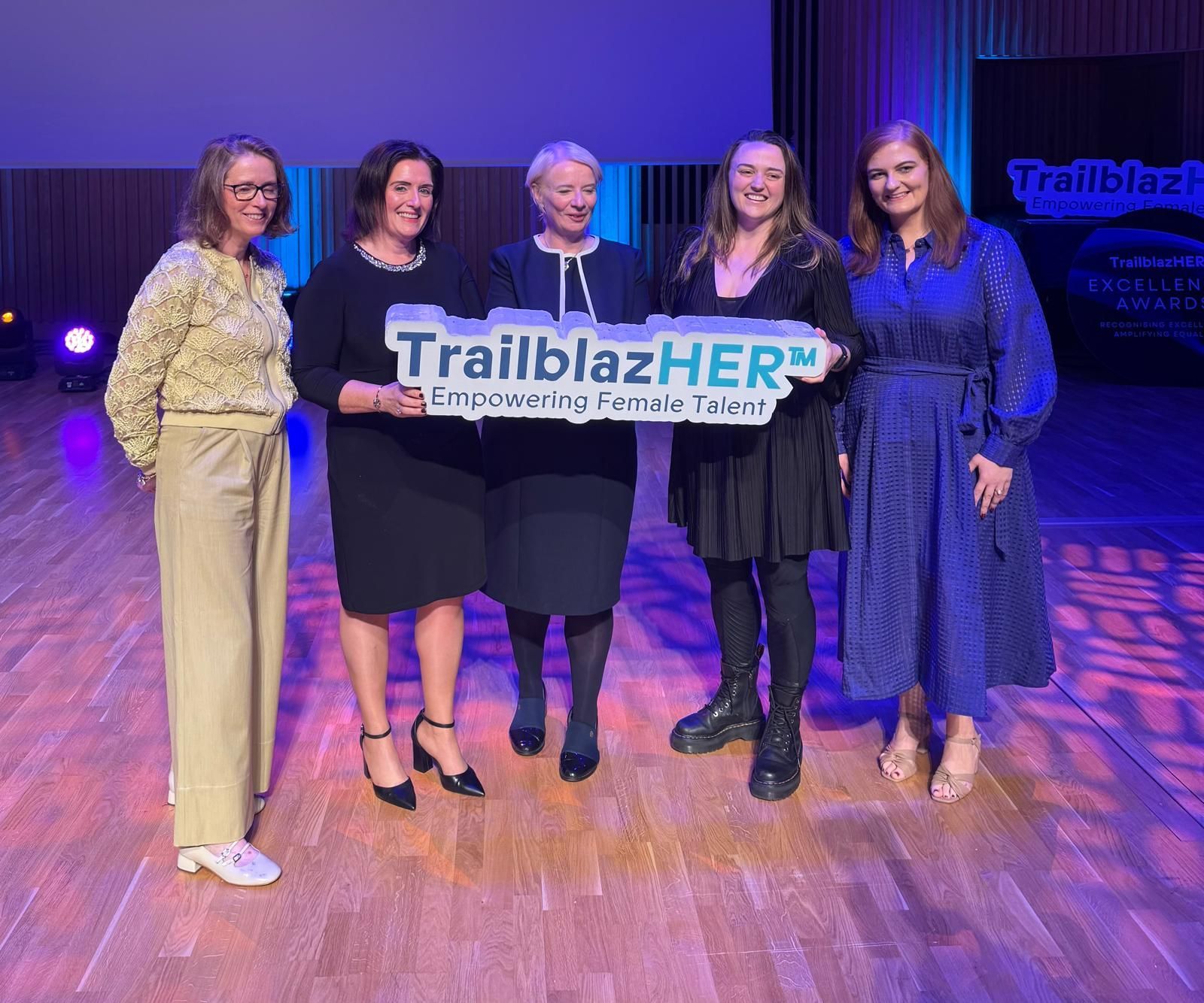Five people on a stage holding a TrailblazHER banner under purple lighting