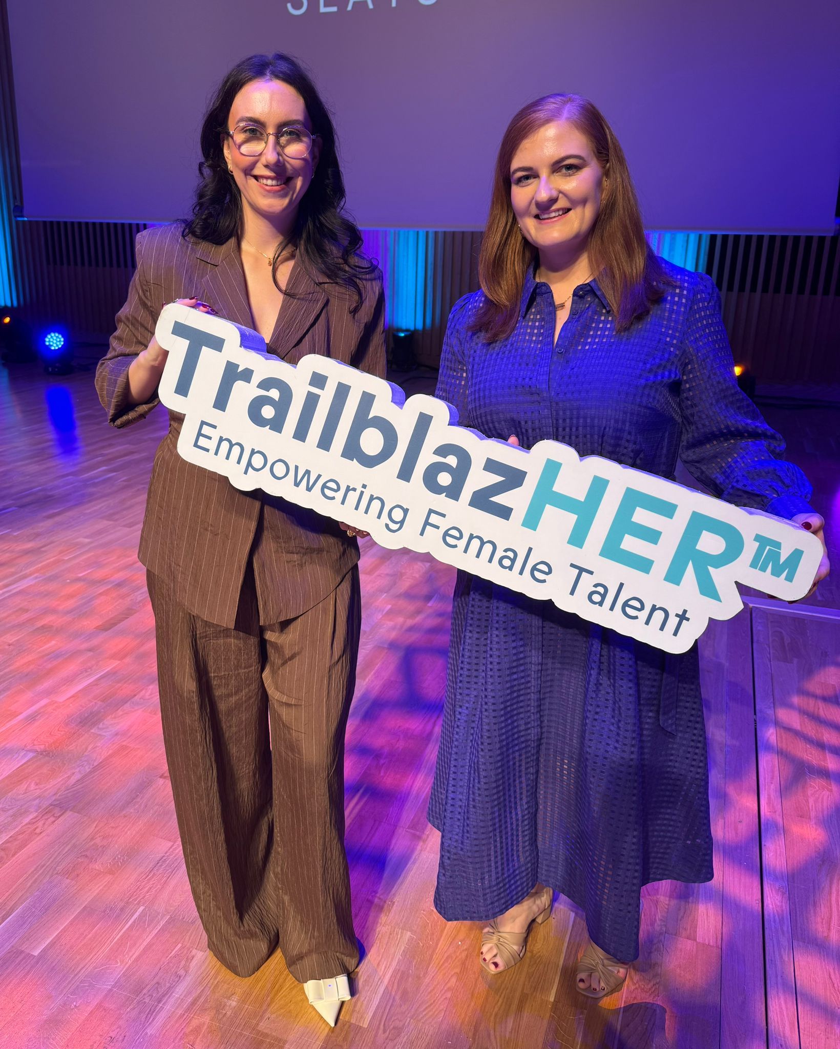 Two women holding a TrafblazHER sign on a lit stage with purple lighting