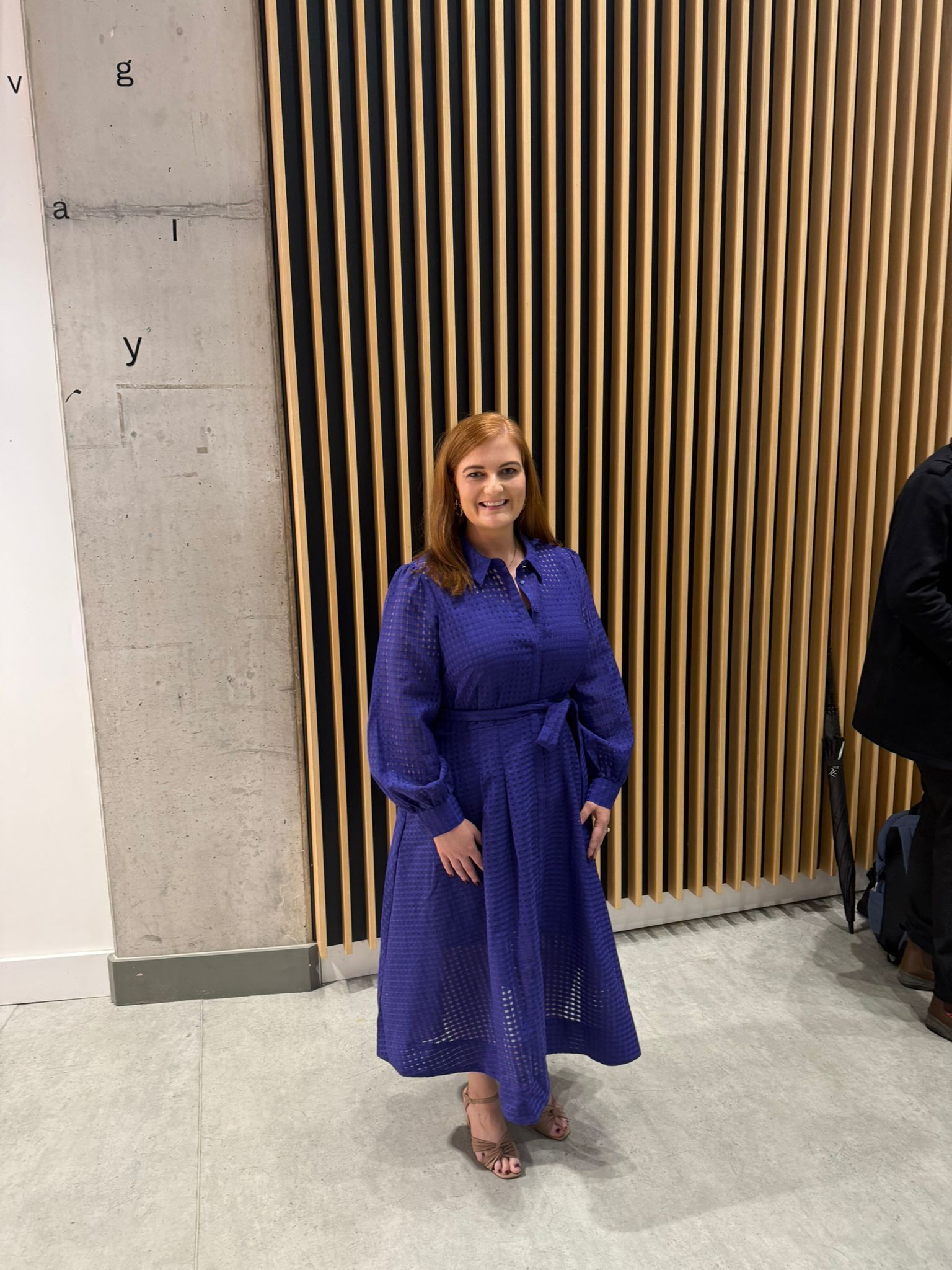 Woman in a purple dress standing indoors beside a wood slat wall