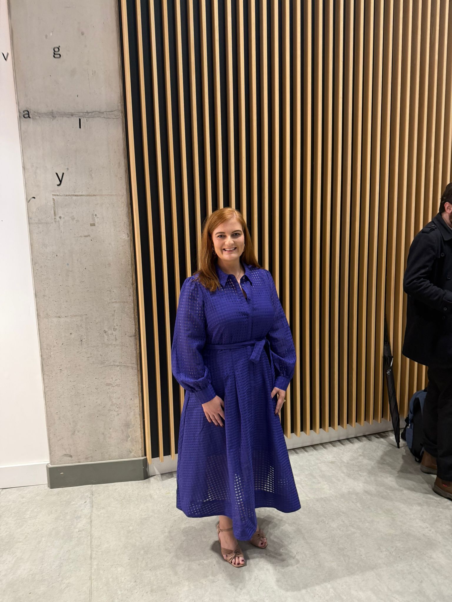 Person in a blue dress standing indoors beside a wall and wooden slats.