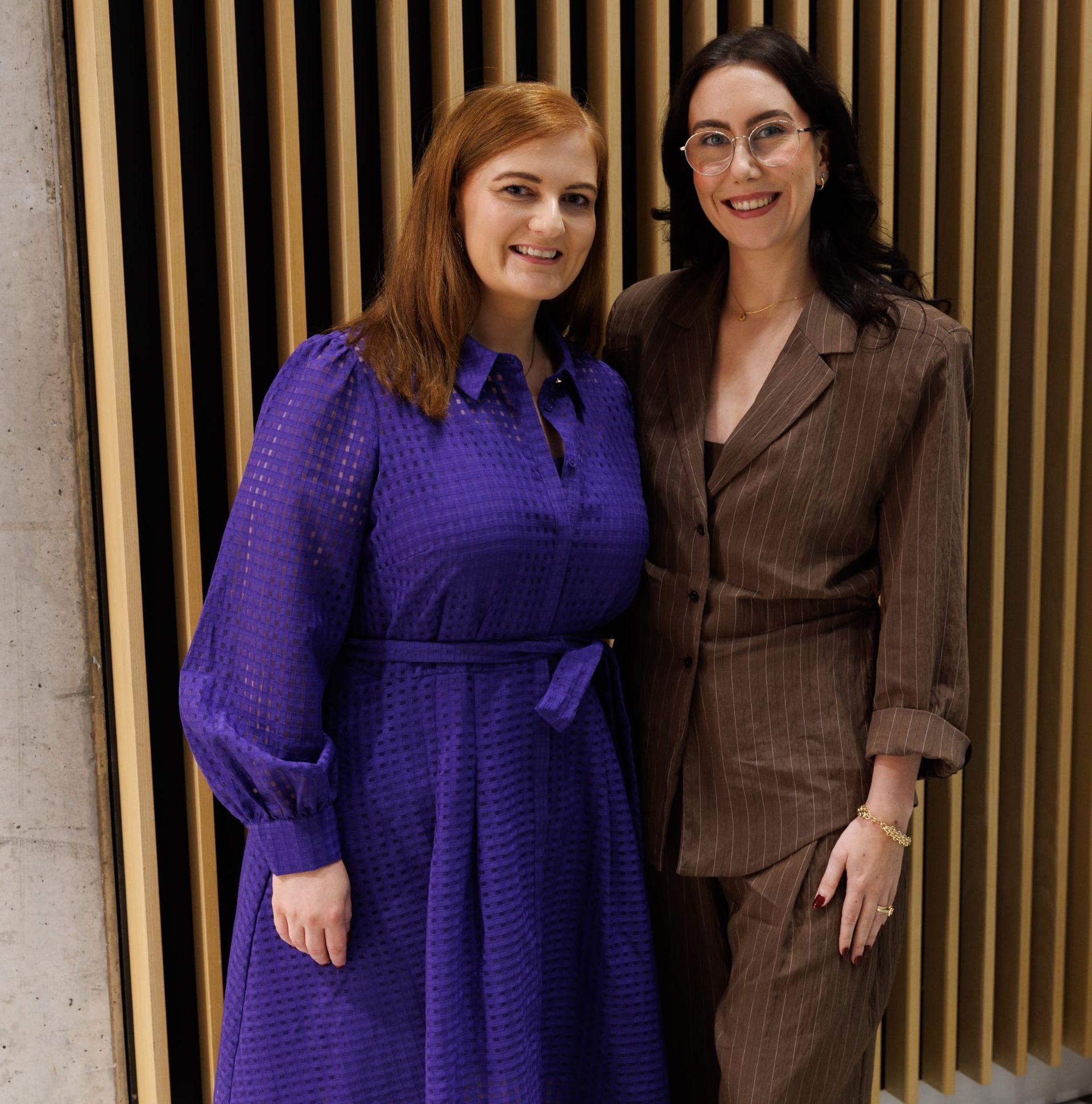 Two women posing together, one in a purple dress and one in a brown suit, against a wood-slatted wall
