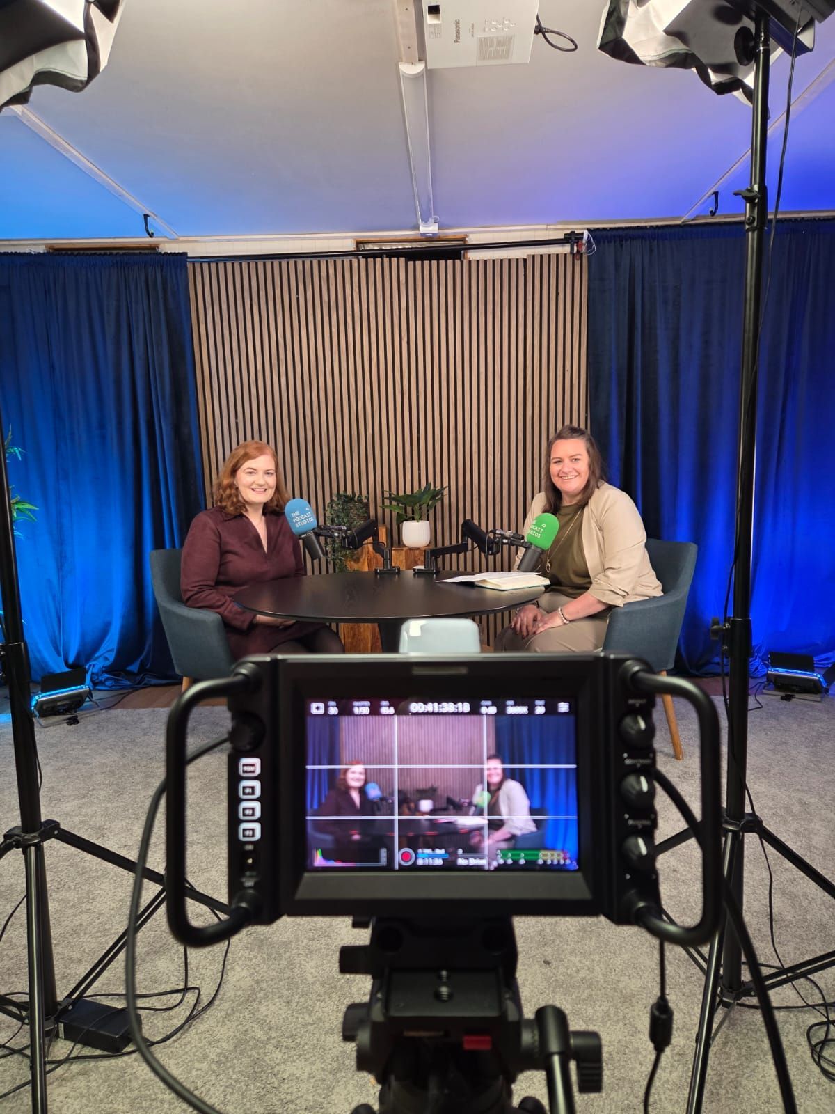 Two people seated at a round table in a studio, seen through a camera monitor and tripods.