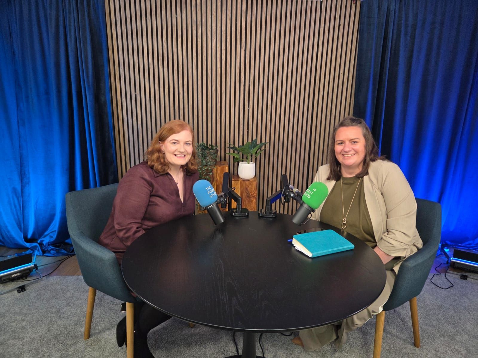Two women seated at a round table with microphones in a studio setting, smiling at the camera.