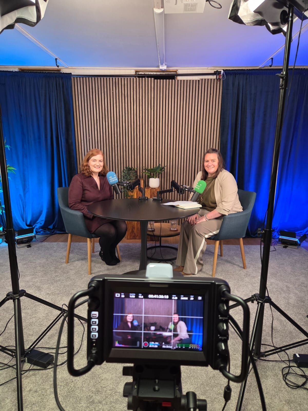 Two people seated at a round table in a studio, filmed on a camera monitor in the foreground