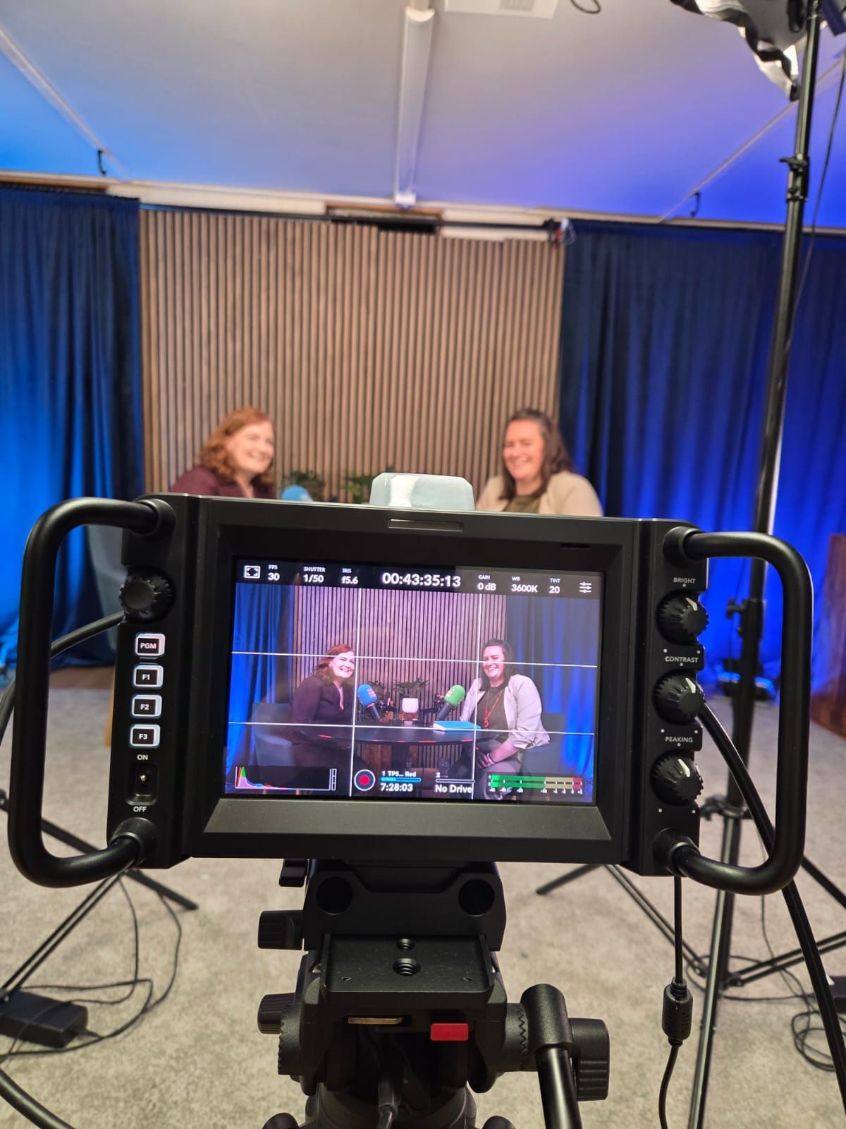 Camera screen showing two women talking at a table on a stage with blue curtains.