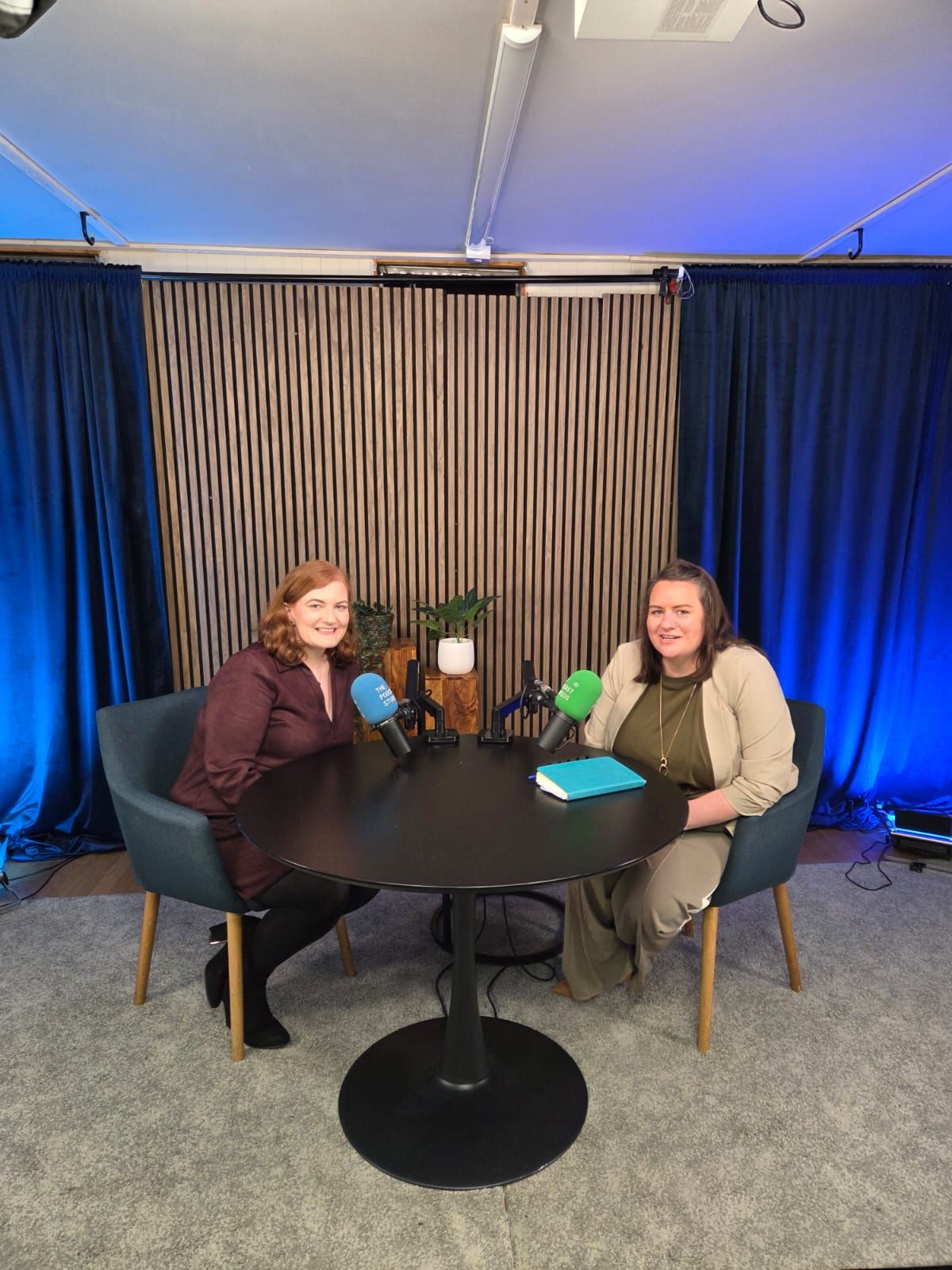 Two people seated at a round table in a studio with microphones, speaking to each other.