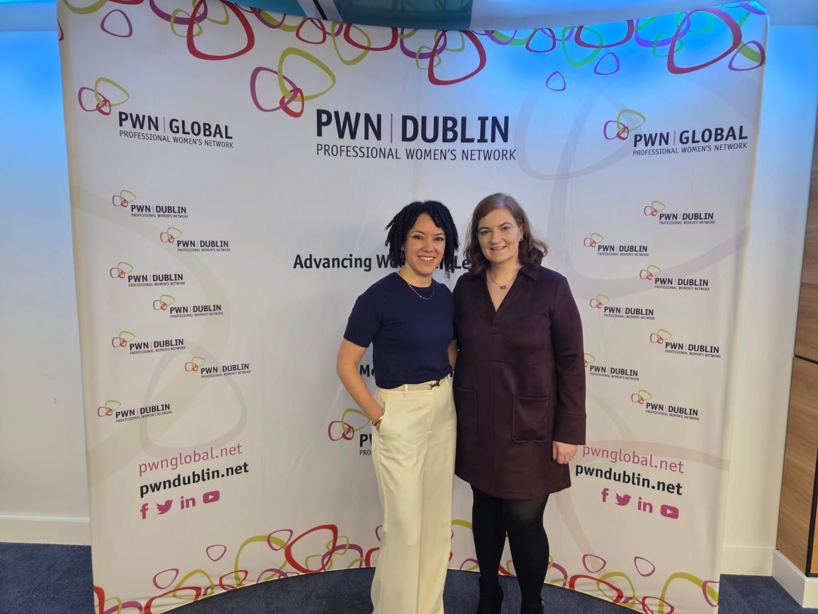 Two people standing smiling in front of a PWN Dublin event backdrop, one in navy and cream, one in burgundy.