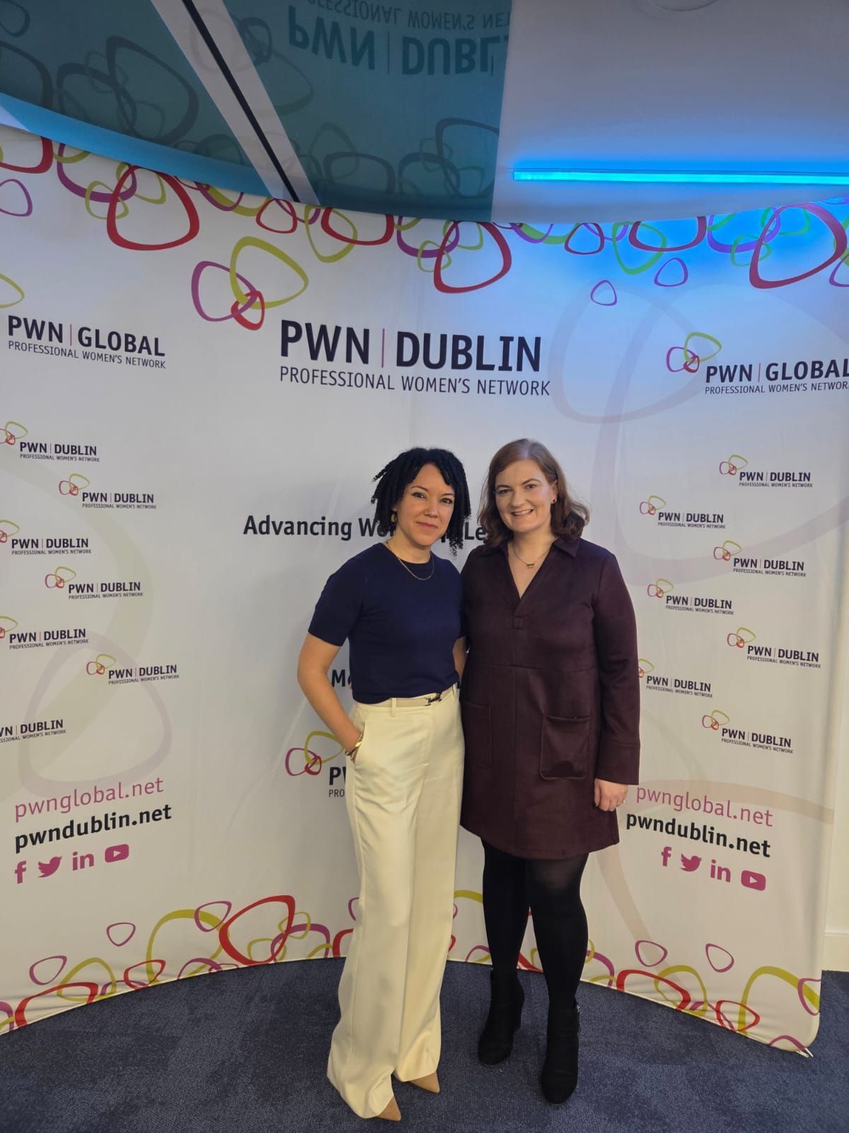 Two women posing at the PWN Dublin event backdrop, smiling in front of a logo wall