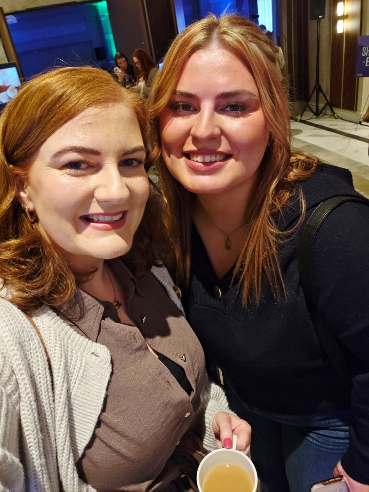 Two smiling people posing indoors, one holding a drink, with warm lighting and event decor in the background