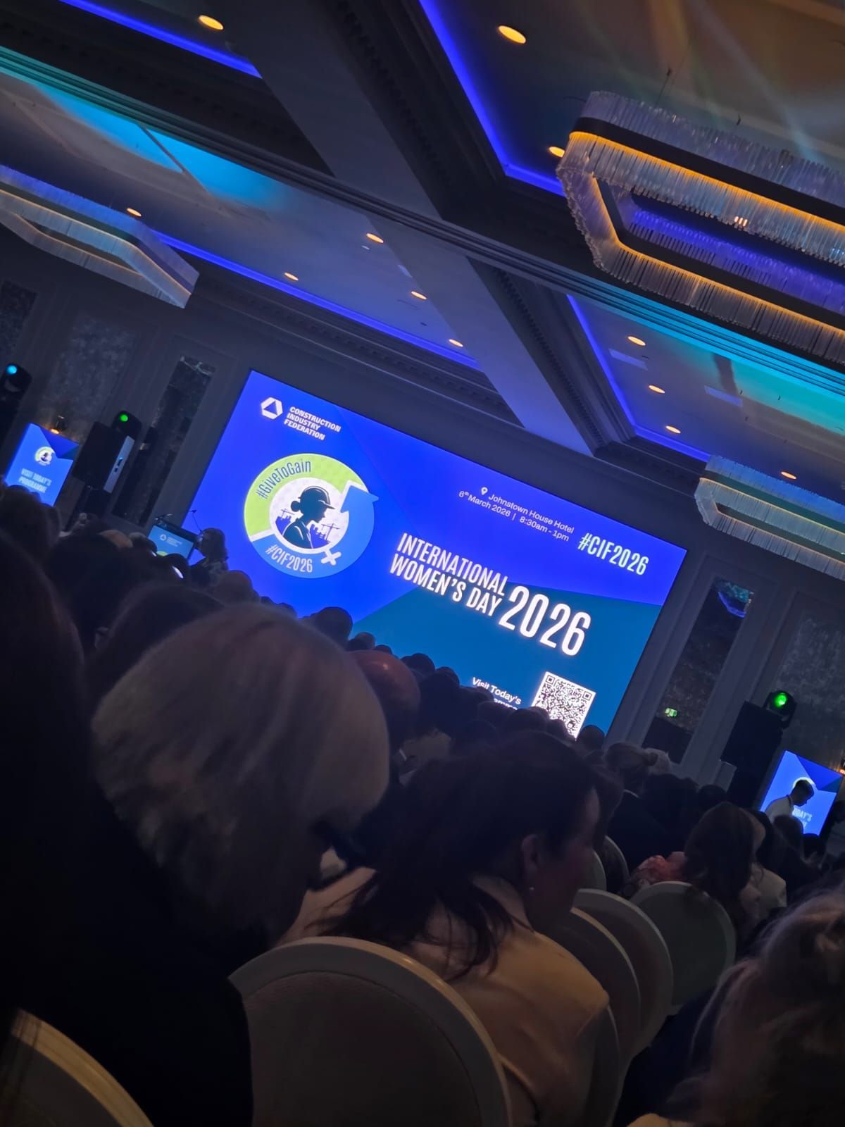 Audience in a dim conference hall watching a blue presentation screen with “2026” visible.