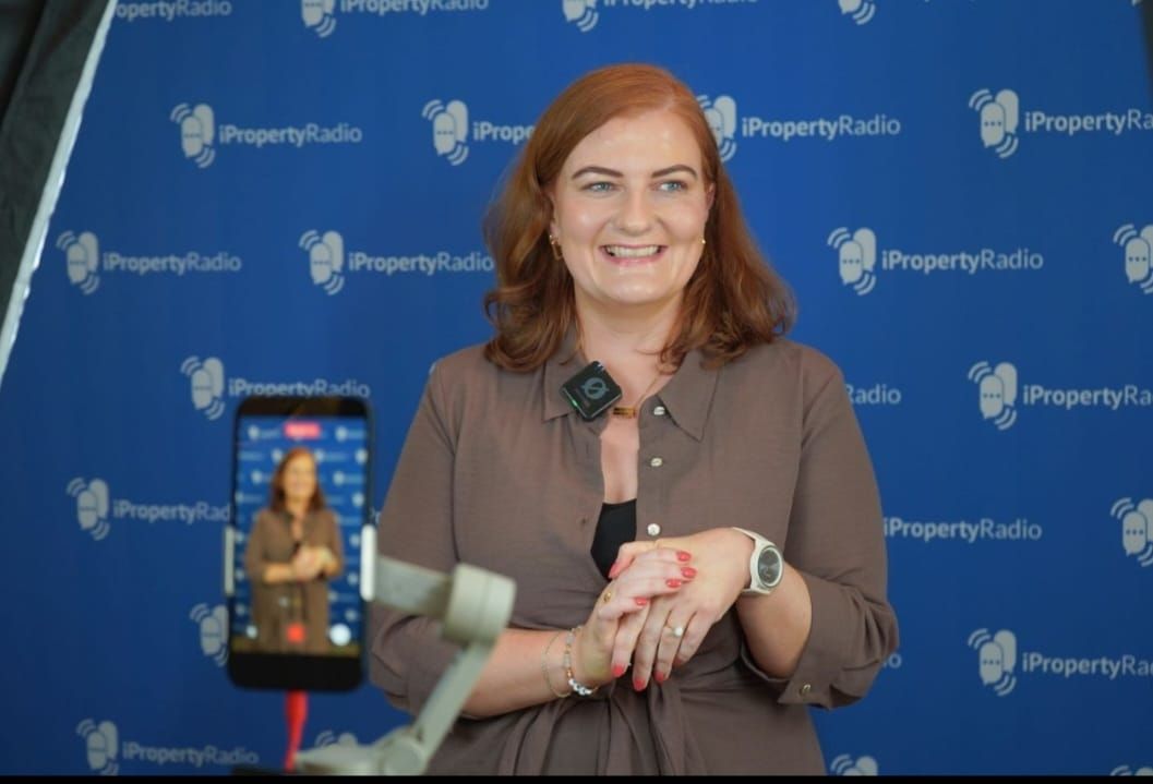 Woman speaking at PropertyGuru event, smiling in front of blue logo backdrop while being filmed by a smartphone.