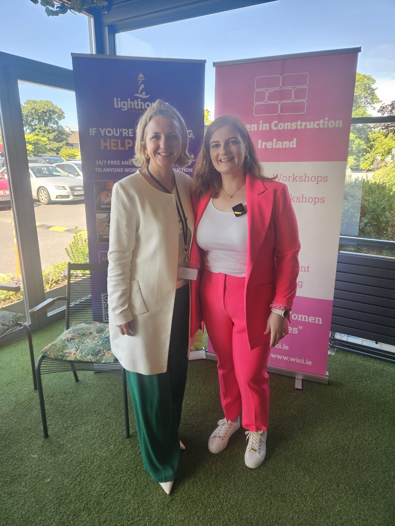 Two women standing outdoors beside event banners, smiling and posing together.