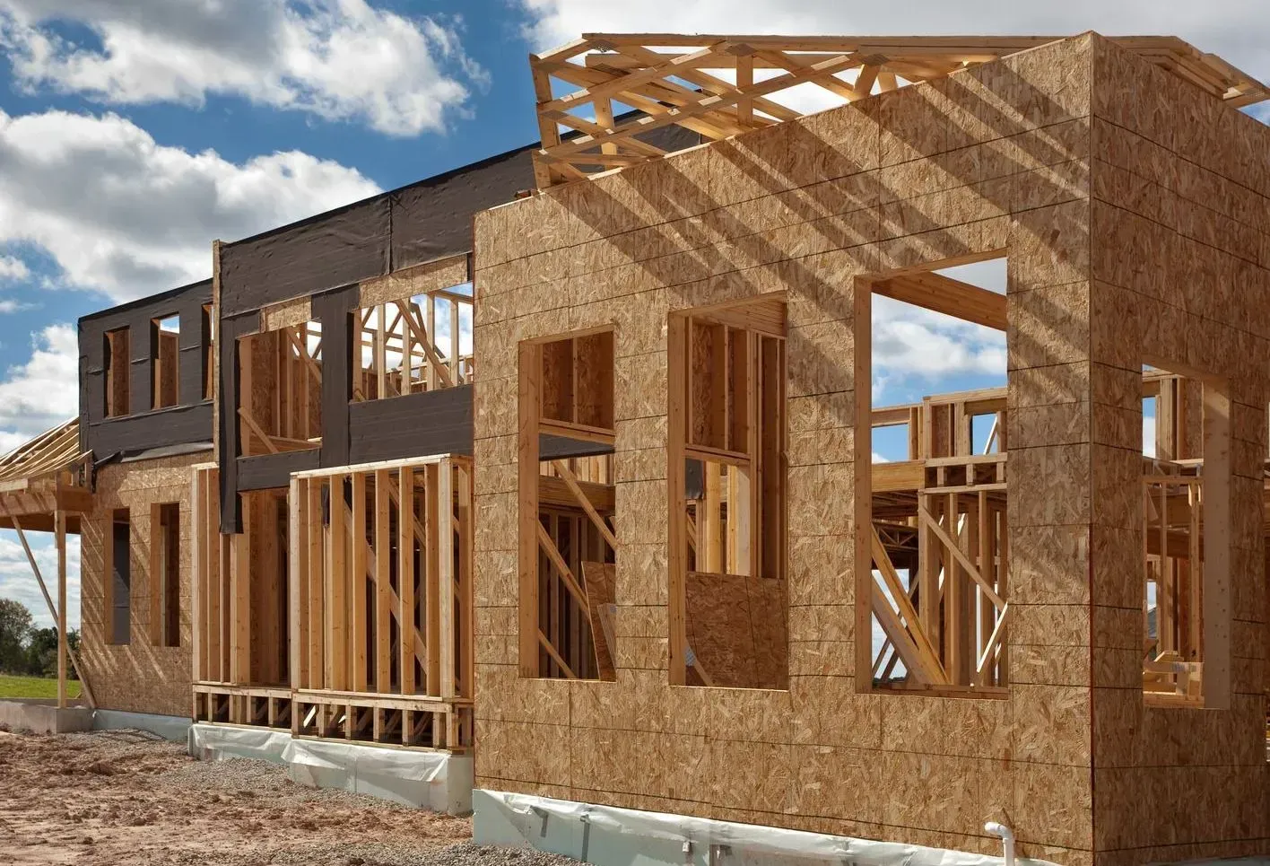 Construction of a two-story wood-framed house with exposed beams, windows, and OSB sheathing against a blue sky.