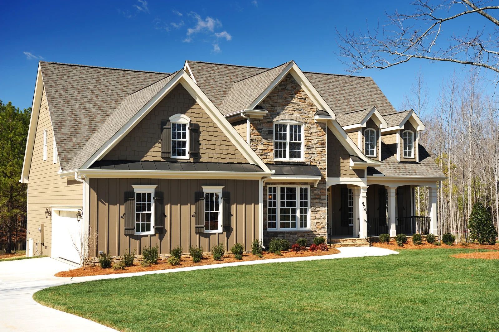 Large beige suburban house with dormer windows, a front porch, and a green lawn under a blue sky