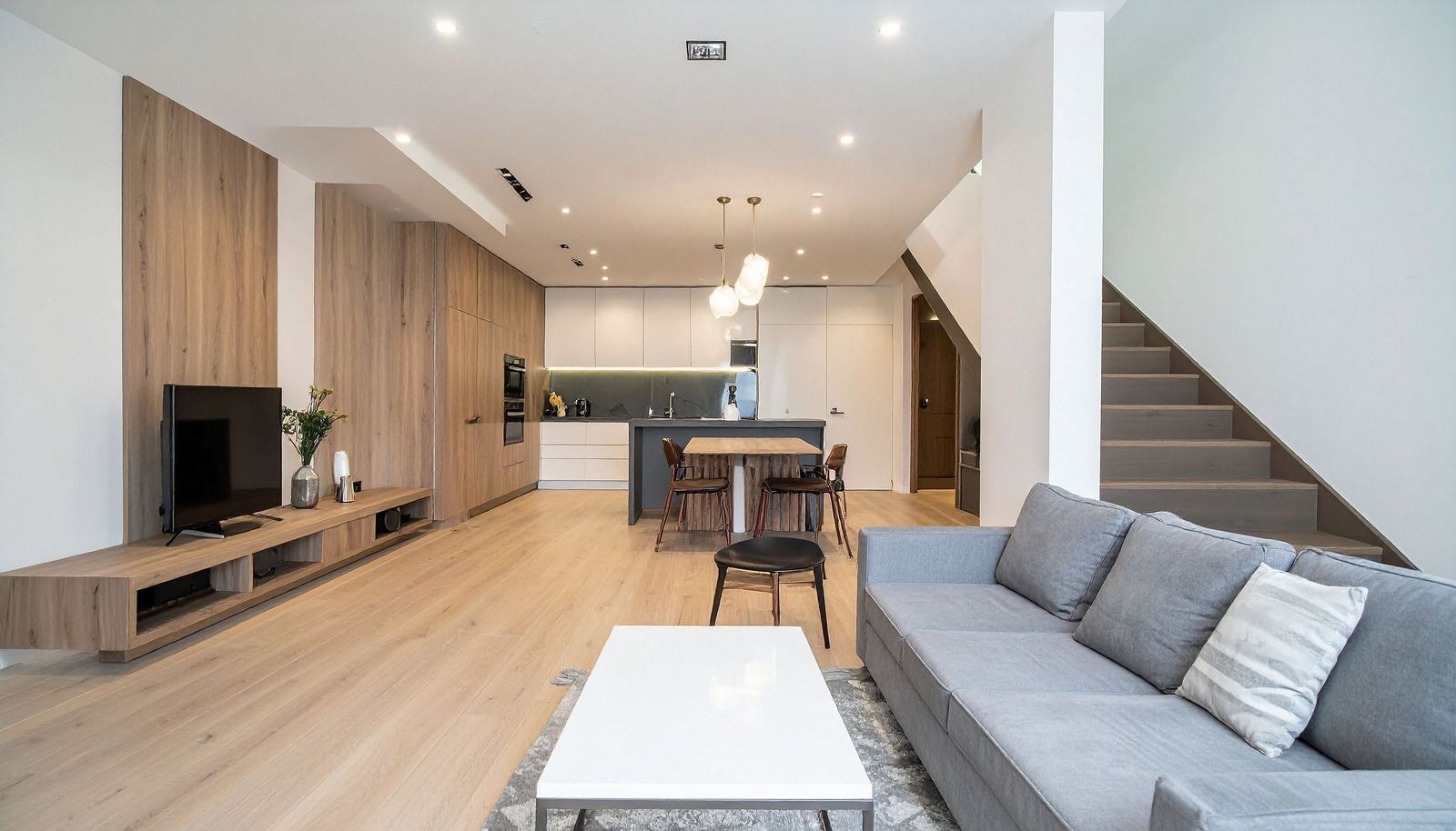 Modern open-plan living room with light wood floors, gray sofa, staircase, dining table, and kitchen area