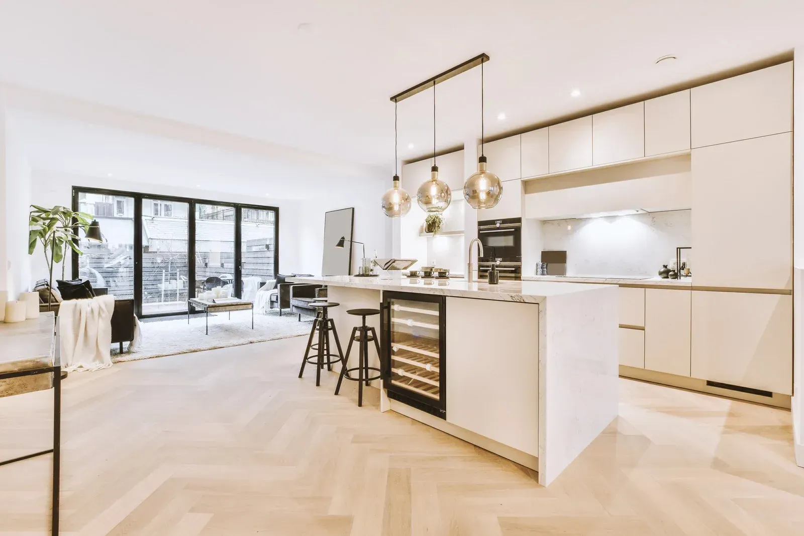 Modern white kitchen with island, wine cooler, and pendant lights. Glass doors open to a patio.