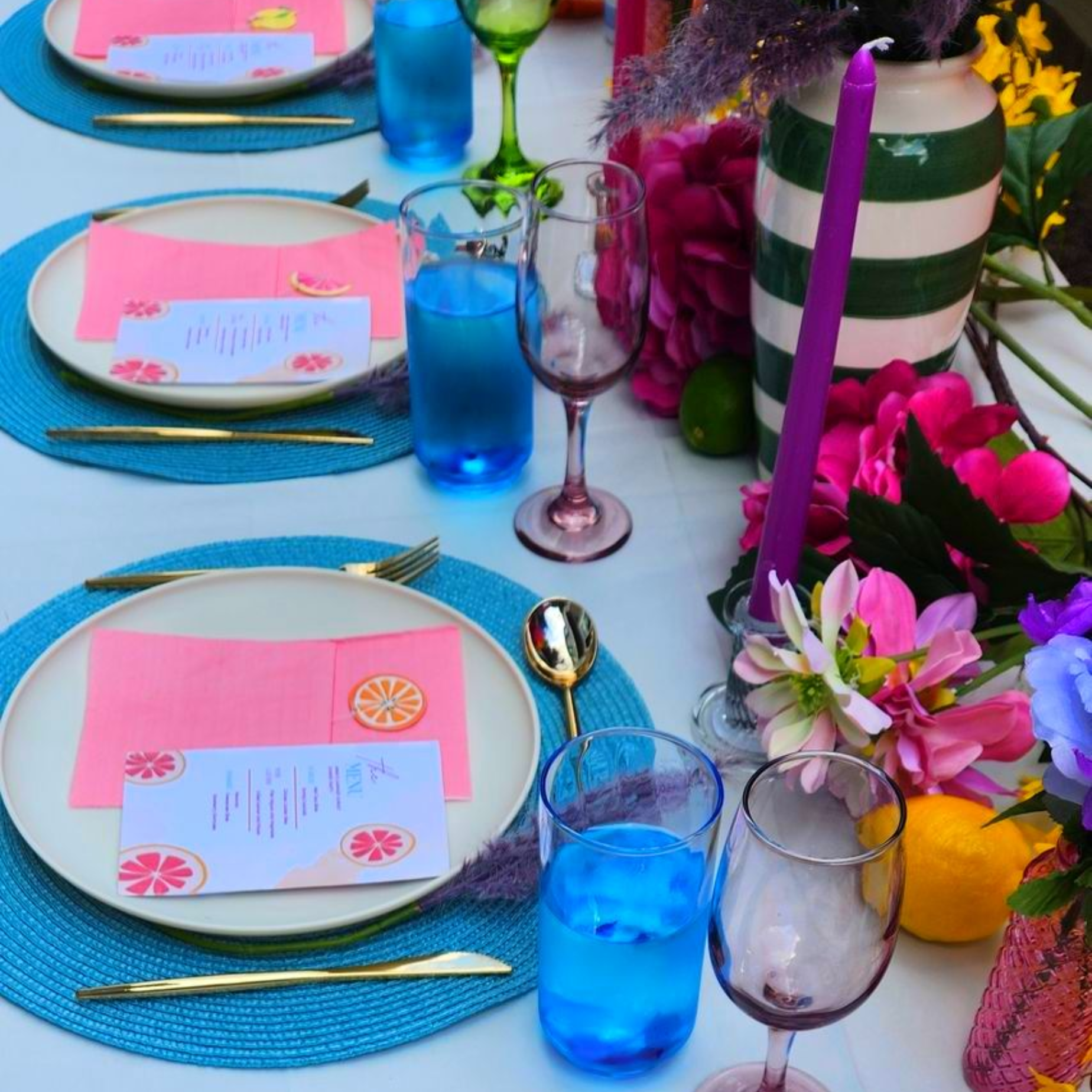 Brightly set outdoor table with cream plates, pink napkins, citrus menus,, candles, and colorful glassware for a celebration