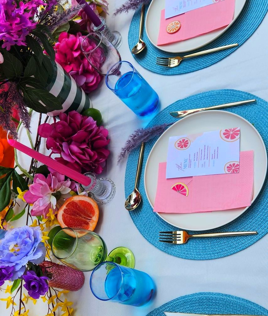 Bright floral table setting with cream plates, pink napkins, citrus menus, glassware, and colorful flowers on a white tablecloth