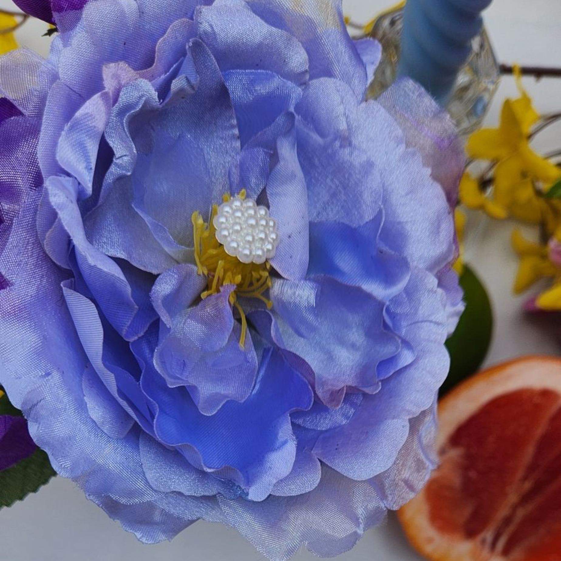 Close-up of a purple-blue flower with a white center, with yellow flowers and an orange slice in the background