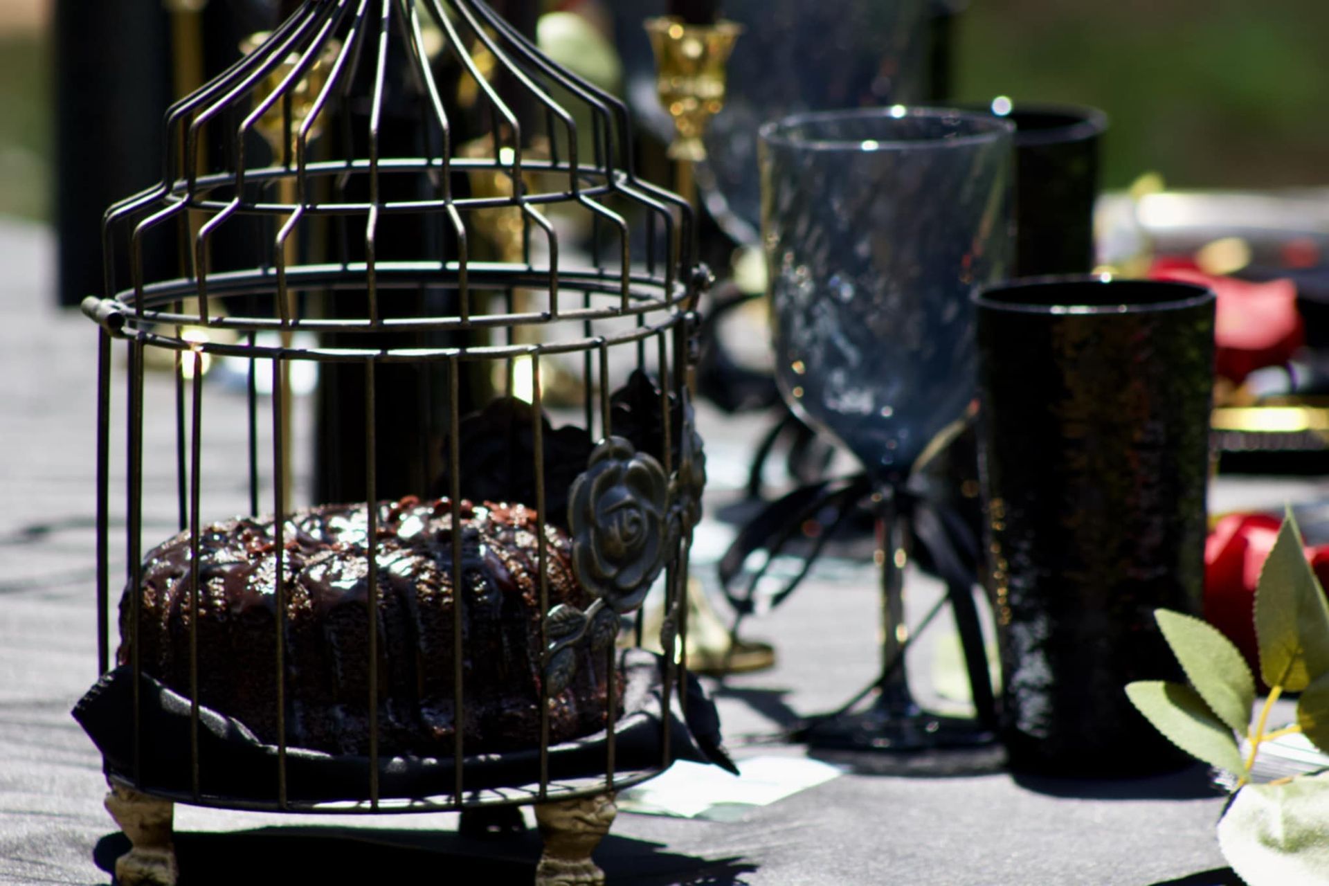Decorative birdcage with a chocolate cake on a table, beside black glassware and a small flower arrangement