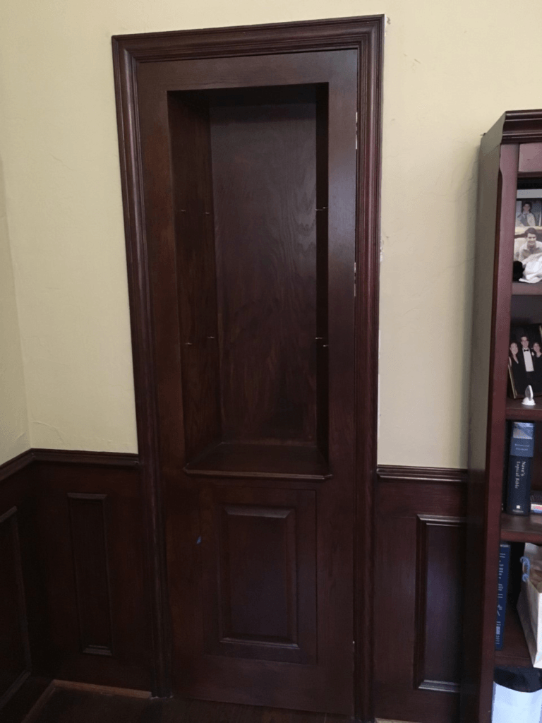 Dark wooden interior door with paneling, set in a beige room with a bookshelf on the right.