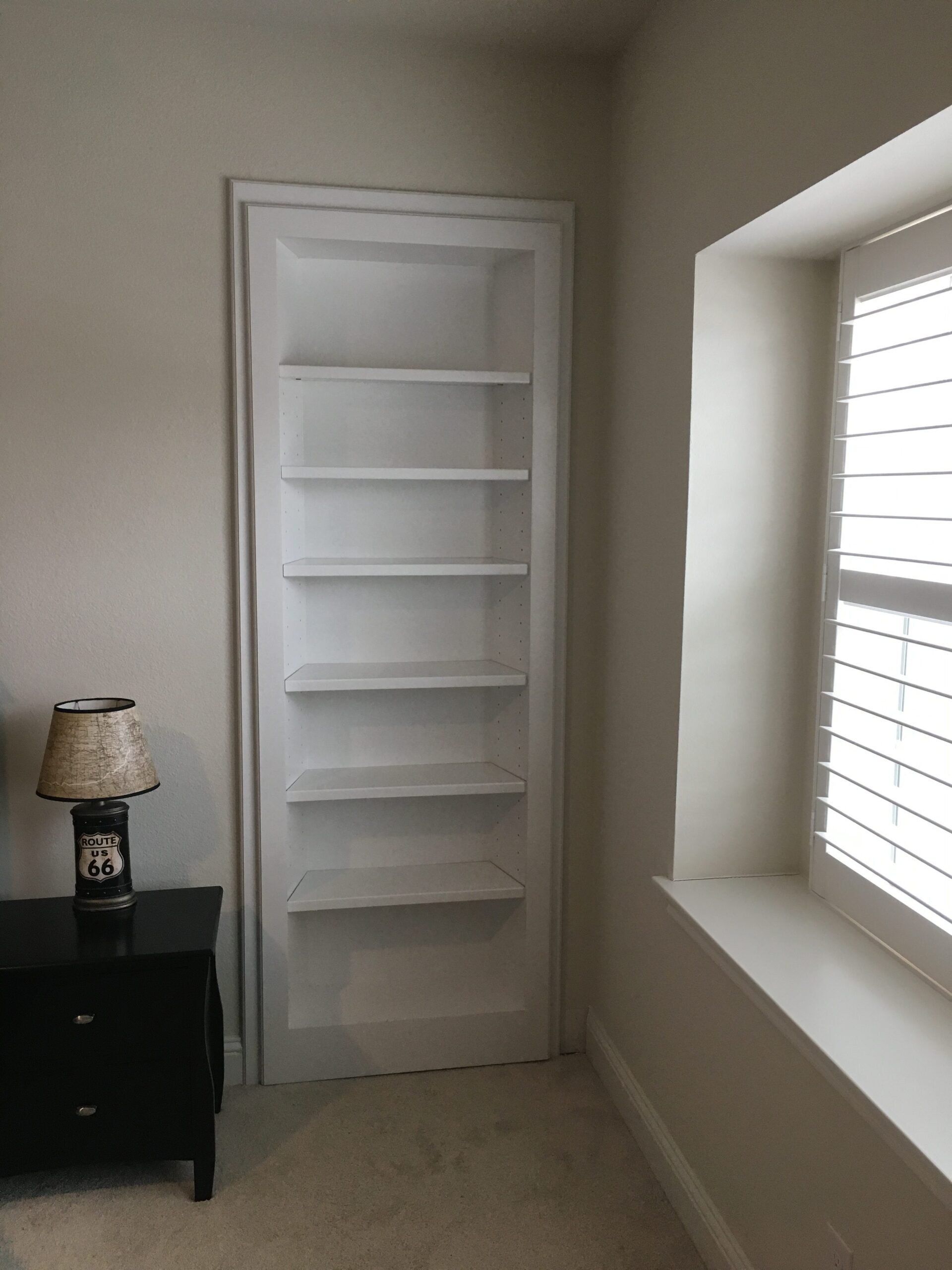 Empty white built-in shelving nook beside a window in a beige room.