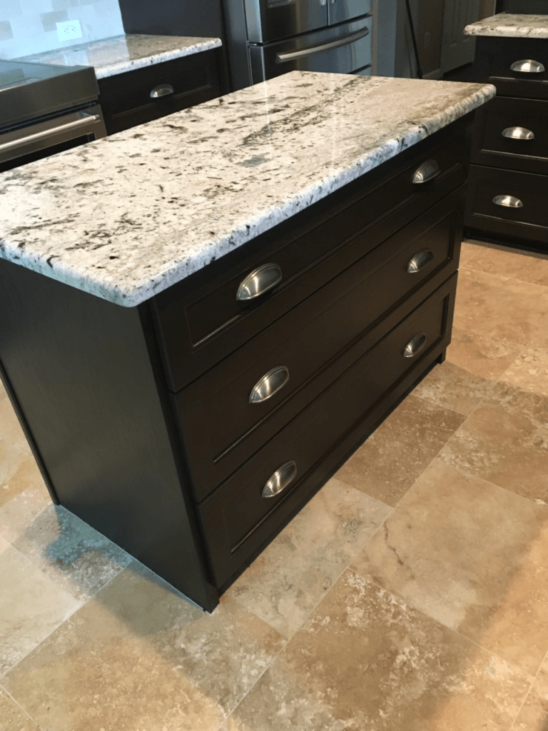 Black kitchen island with a speckled granite countertop and three drawers on a tiled floor