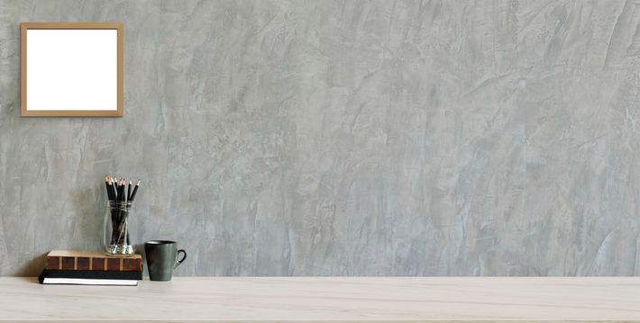Una stampa artistica incorniciata è appesa sopra una scrivania con matite, libri e una tazza contro un muro di cemento grigio.
